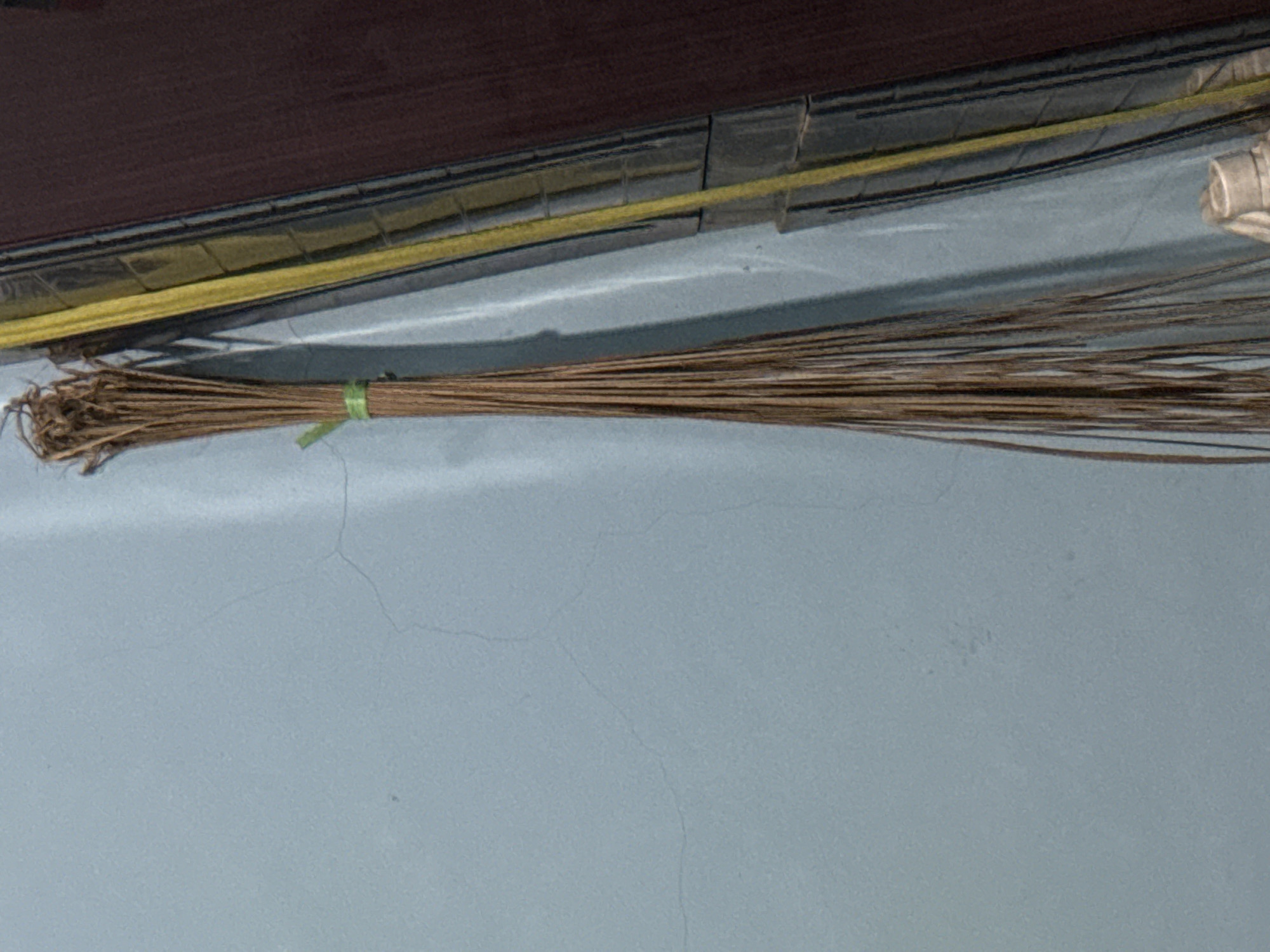 A broom made of dried grass stalks, bound by a green string, hangs vertically against a light blue wall. To the right of the broom is a dark reddish-brown object with a vertical yellow stripe. The broom is positioned in the foreground. The background consists of a textured blue wall with faint cracks. The scene appears to be indoors, possibly in a room or alleyway.