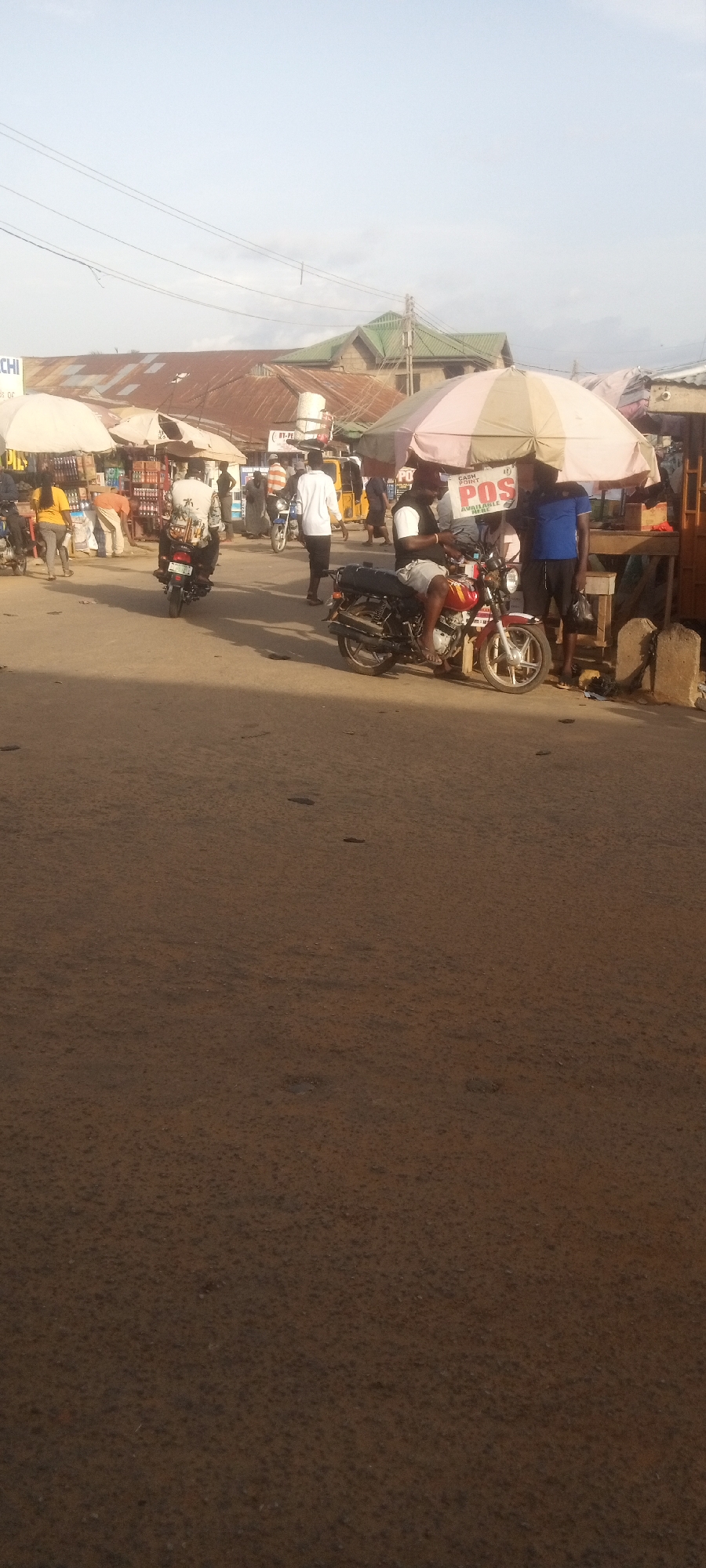 The image depicts a street scene in Narayi, Nigeria. The foreground shows a large expanse of dirt road. In the middle ground, people are present, with at least three motorcycles in view. One motorcycle is parked, and a man sits on it while interacting with two other men standing nearby under a pink and white striped umbrella with "POS" printed on it. Other motorcycles and pedestrians are moving in the background. Temporary market stalls are set up along the side of the road, covered by umbrellas. Buildings with corrugated iron roofs are visible in the background. Overhead electrical wires stretch across the sky.