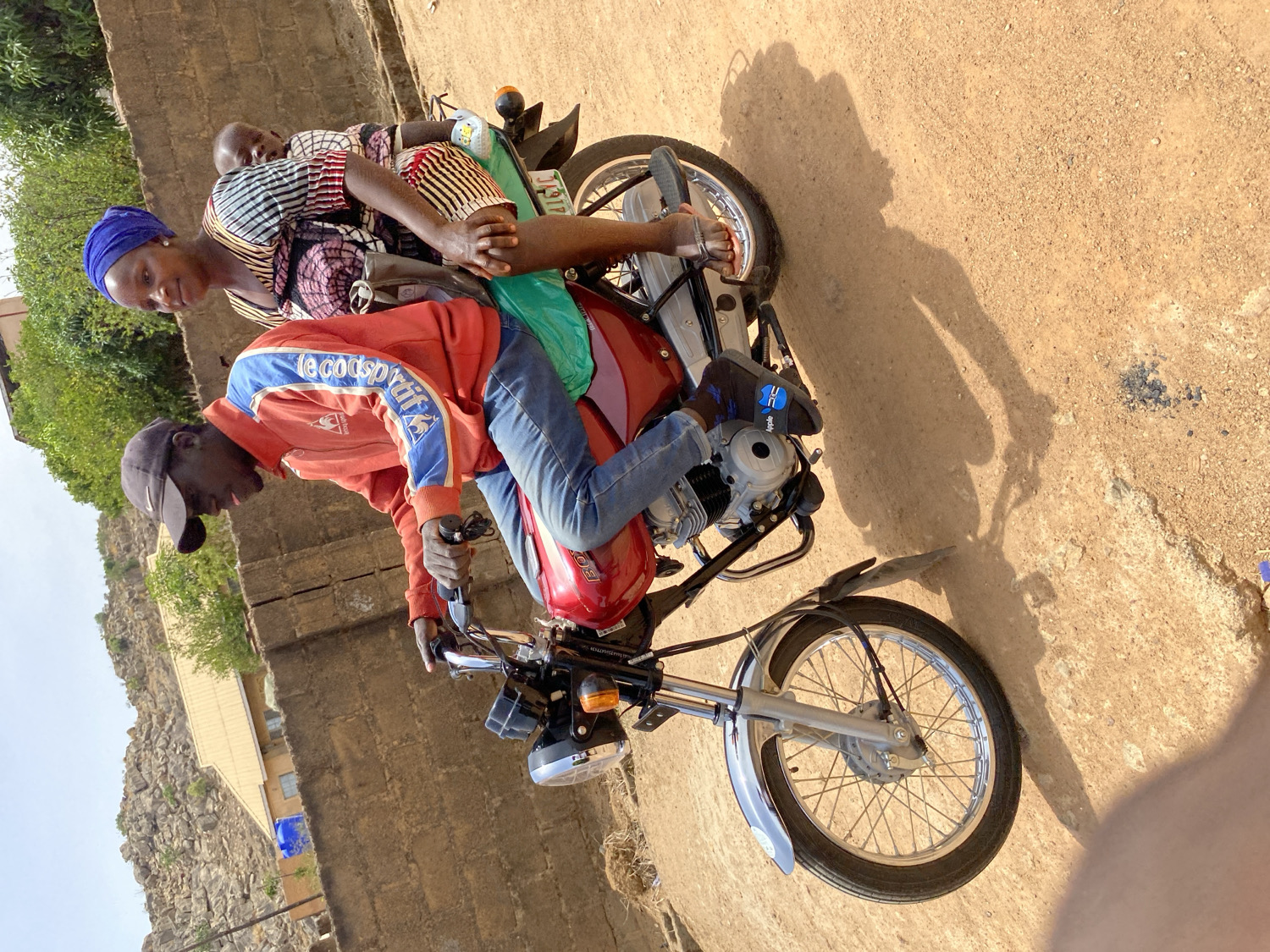 The image, correctly oriented, displays three individuals on a red motorcycle on an unpaved road, bathed in bright sunlight. The scene is consistent with a rural or semi-urban setting, likely Barakin Akawo, Nigeria.
The foreground features a man, who appears to be the driver, seated on the motorcycle. He wears a black baseball cap, an orange and blue long-sleeved jacket with "Le Coq Sportif" visible on the sleeve, blue jeans, and dark sneakers adorned with an "Apple" logo. He is leaning forward, holding the handlebars. Behind him sits a woman, identifiable by her blue headscarf and a multi-colored striped and patterned dress. She looks towards the viewer with a slight smile. A child, mostly obscured by the woman, is seated at the very back. A green plastic bag is draped over the rear of the motorcycle's seat.
The motorcycle is red with chrome details on the front fender and silver spokes on the wheels. Faint lettering on the fuel tank suggests a brand, possibly "HONDA." The rear license plate holder clearly shows "KANO," indicating Kano State in Nigeria, along with partial alphanumeric characters.
The ground is a light brown, sandy or dirt surface, with prominent, long shadows cast by the people and the motorcycle, suggesting a sunny day, likely in the mid-morning or early afternoon. In the background, there are low-rise buildings constructed from light-colored materials, sparse green vegetation, and rocky, hilly terrain under a clear sky. The image captures a routine moment of local transportation.