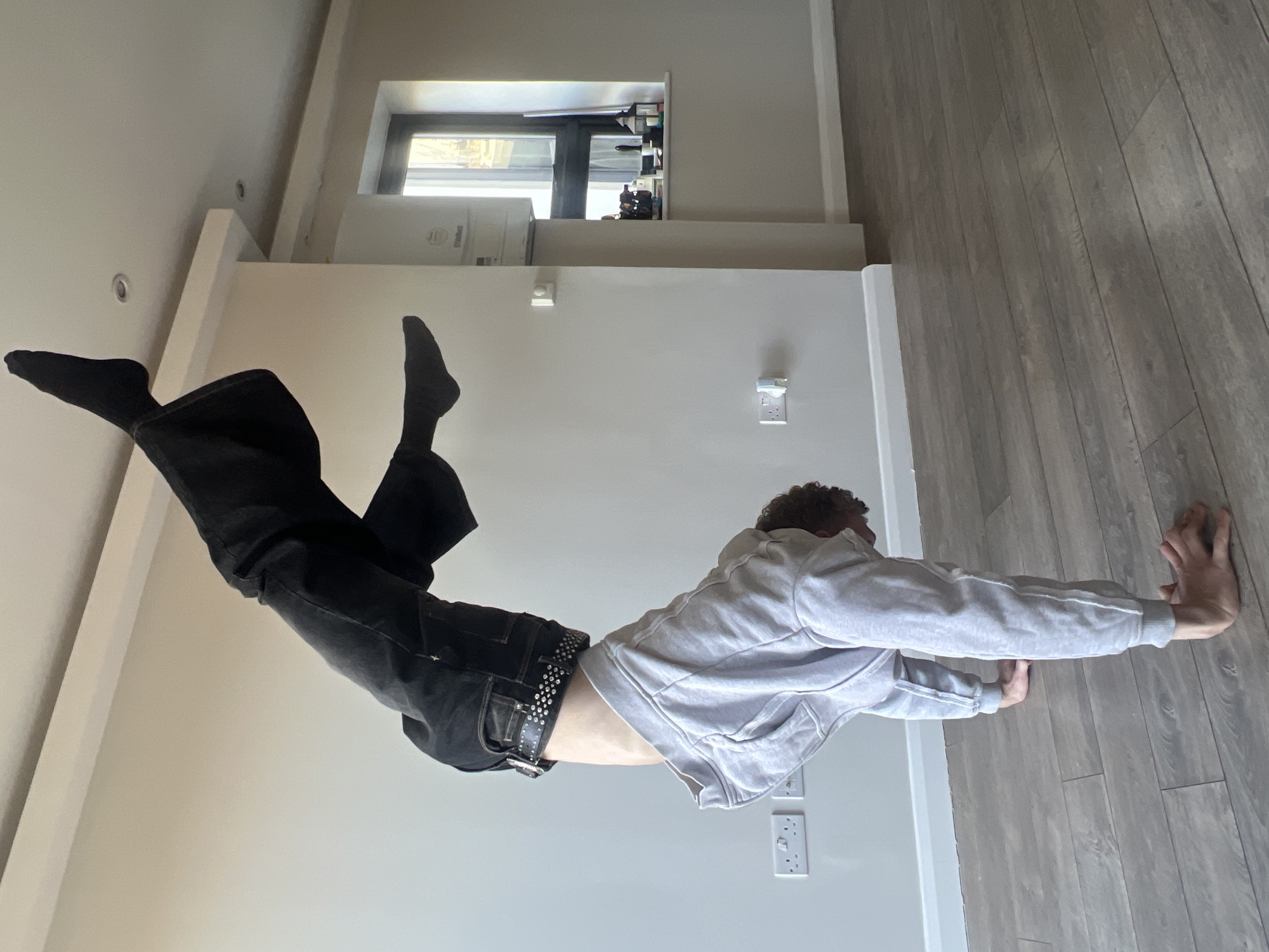 A person wearing black socks, black trousers, a belt, and a light gray hoodie is performing a yoga pose on a hardwood floor. The person's hands are pressed against the floor while their legs are elevated in the air. The background includes a white wall, electrical outlets, and a window. The location appears to be indoors, possibly a residence, and it is unknown if it is in the city of London, United Kingdom.