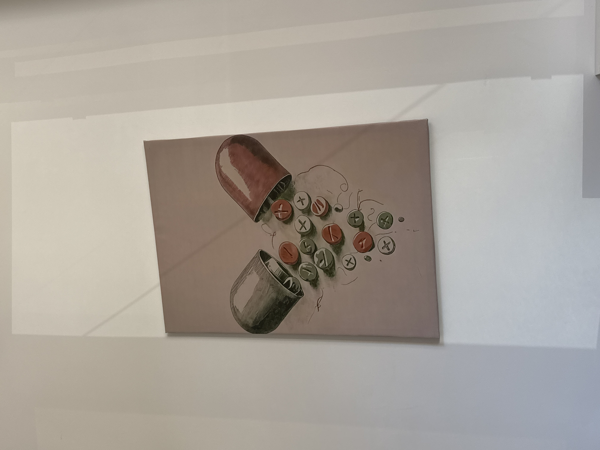 The image shows a framed artwork depicting an opened pill capsule on a white wall. The pill's two halves, colored in reddish-brown and grey, are positioned as if spilling out circular tablets. These tablets are decorated with mathematical symbols like +, -, x, and ÷. The artwork has a pale pink background. The setting is an interior space, likely in Yerevan, Armenia. No people are present in the frame.
