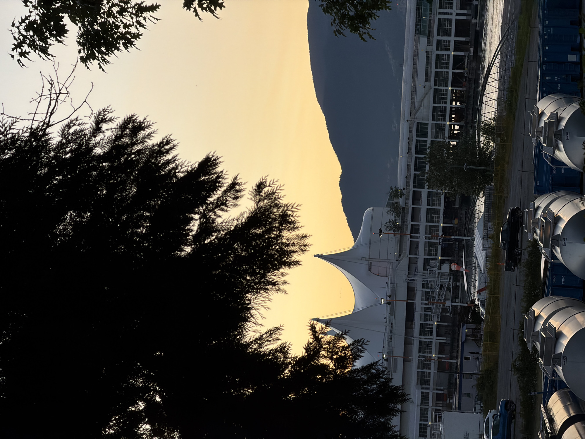 The image shows a dusk scene at Canada Place in Vancouver, Canada. In the foreground, several large, cylindrical tanks with metallic exteriors are visible, some with "POLAR" labeling. Behind the tanks, a road is present with a blue sedan on the left and a black pickup truck on the right. Further back is a large, white building with a distinctive tent-like roof structure, characteristic of Canada Place. The building is multi-story and has numerous windows. A body of water is visible in the middle ground, reflecting the light. Beyond the building and water, a dark, silhouetted mountain range dominates the background. The sky above is a gradient of pale yellow and orange, indicative of sunset. Dark, silhouetted foliage from trees frames the top and sides of the image.
