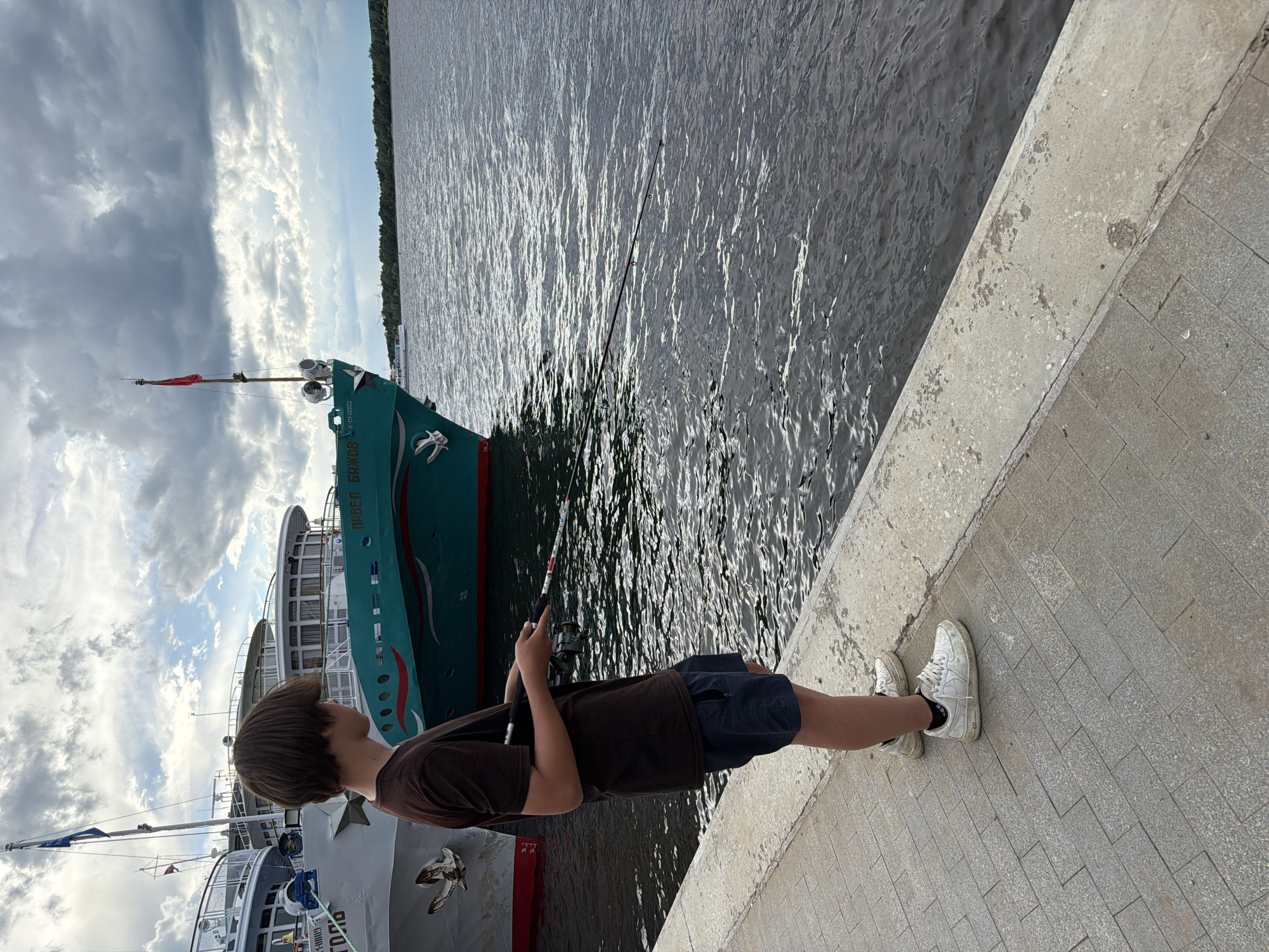The image shows a young person fishing from a concrete embankment beside a body of water, likely a river, in Perm, Russia. The person, facing away from the camera, is holding a fishing rod and wearing a brown t-shirt, dark shorts, and white sneakers. The water is rippling, reflecting the light, suggesting it is daytime. In the background, two large ships are docked. The ship closest to the foreground is partially visible, and the larger ship behind it is a teal color with white accents and lettering. The sky above is partly cloudy, with dramatic, textured clouds mixed with patches of blue sky, indicating fair weather with some cloud cover. The embankment is made of concrete and paving stones. The overall scene conveys a sense of calm and leisure activity. Visible text on the teal ship appears to be in Cyrillic, reading "ПАВЕЛ БАЖОВ".