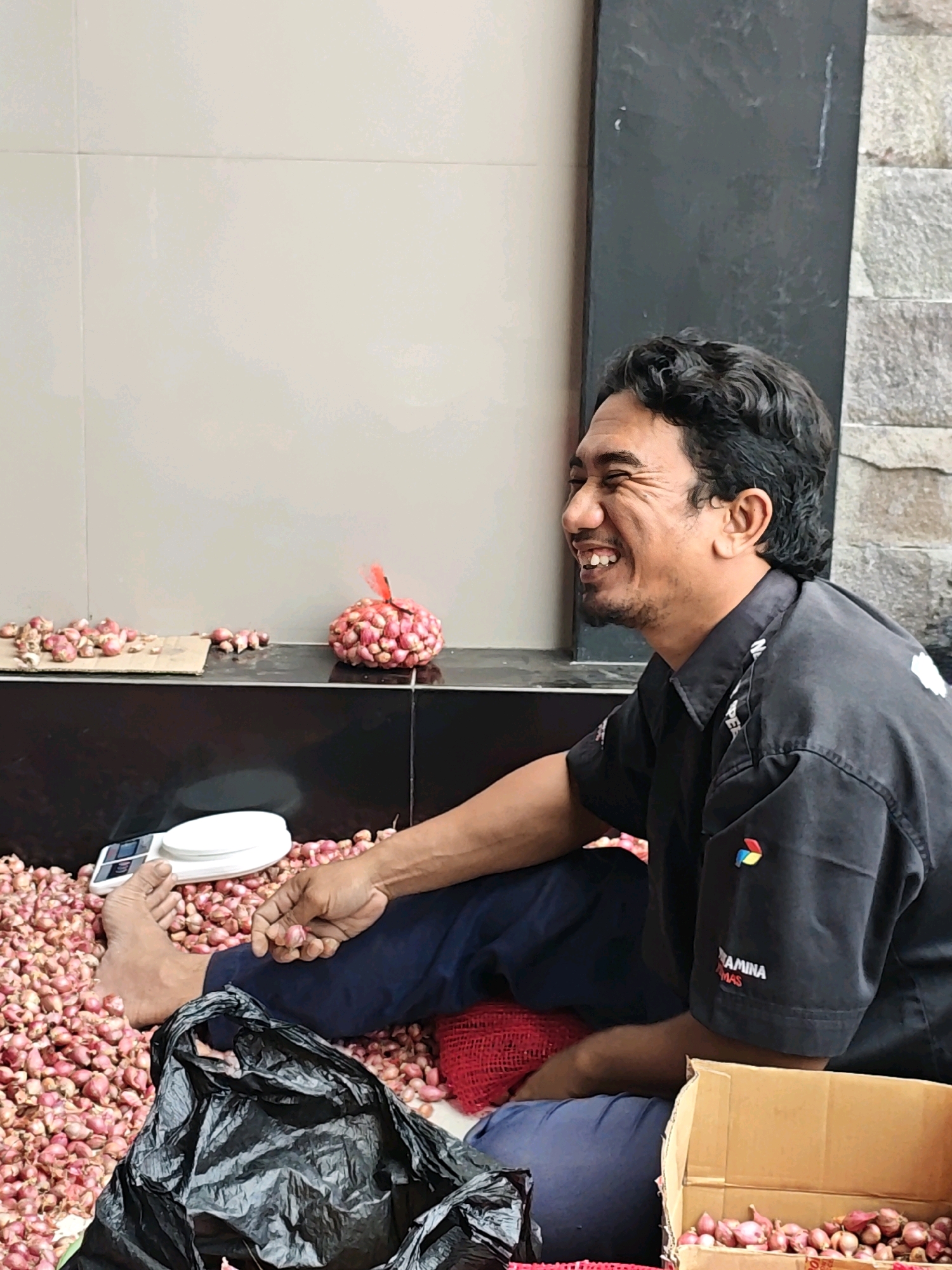 In Ngasem, Indonesia, a man is seated outdoors, surrounded by a large pile of shallots. He is wearing a black uniform shirt with colorful patches on the left sleeve and text that appears to read "PERTAMINA". The man is smiling broadly, his head tilted back slightly, and he is holding a single shallot in his right hand, examining it. In front of him, partially visible, is a white digital scale, and to his left is a black plastic bag. To his right, a cardboard box filled with shallots is positioned on the ground. The background consists of a textured wall made of light-colored stone and a section of smooth, light-colored tile. The ground is also covered with numerous shallots.