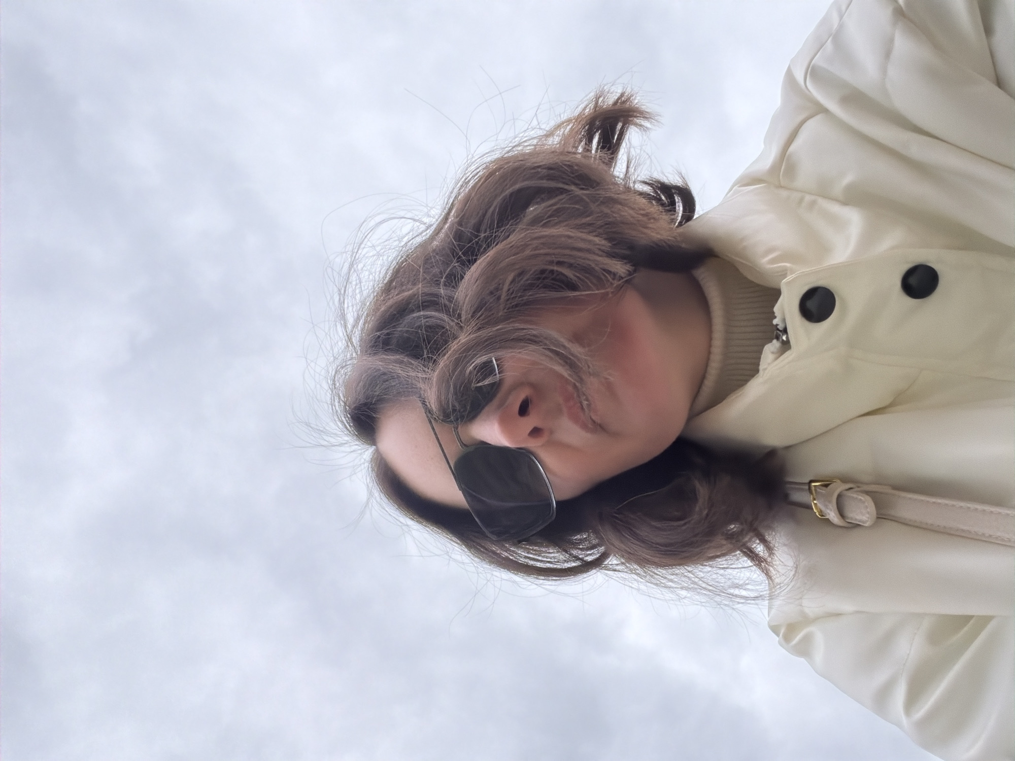 A low-angle shot depicts a woman wearing sunglasses and a cream-colored jacket, set against an overcast sky. The location and city are not identifiable from the image.