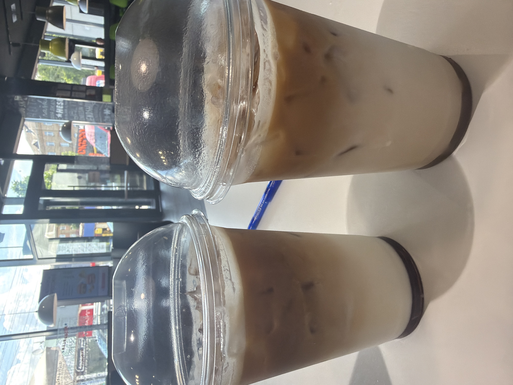 Two iced coffee drinks are on a white table, with a blue pen lying horizontally between them. The drinks are in clear plastic cups with domed lids, filled with ice and a layered mixture of coffee and milk. The background shows a bright interior with large windows looking out onto a city street. Visible through the windows are buildings, trees, and vehicles, suggesting an urban environment. The interior features hanging light fixtures and dark ceiling panels. The overall impression is that of a cafe or coffee shop.