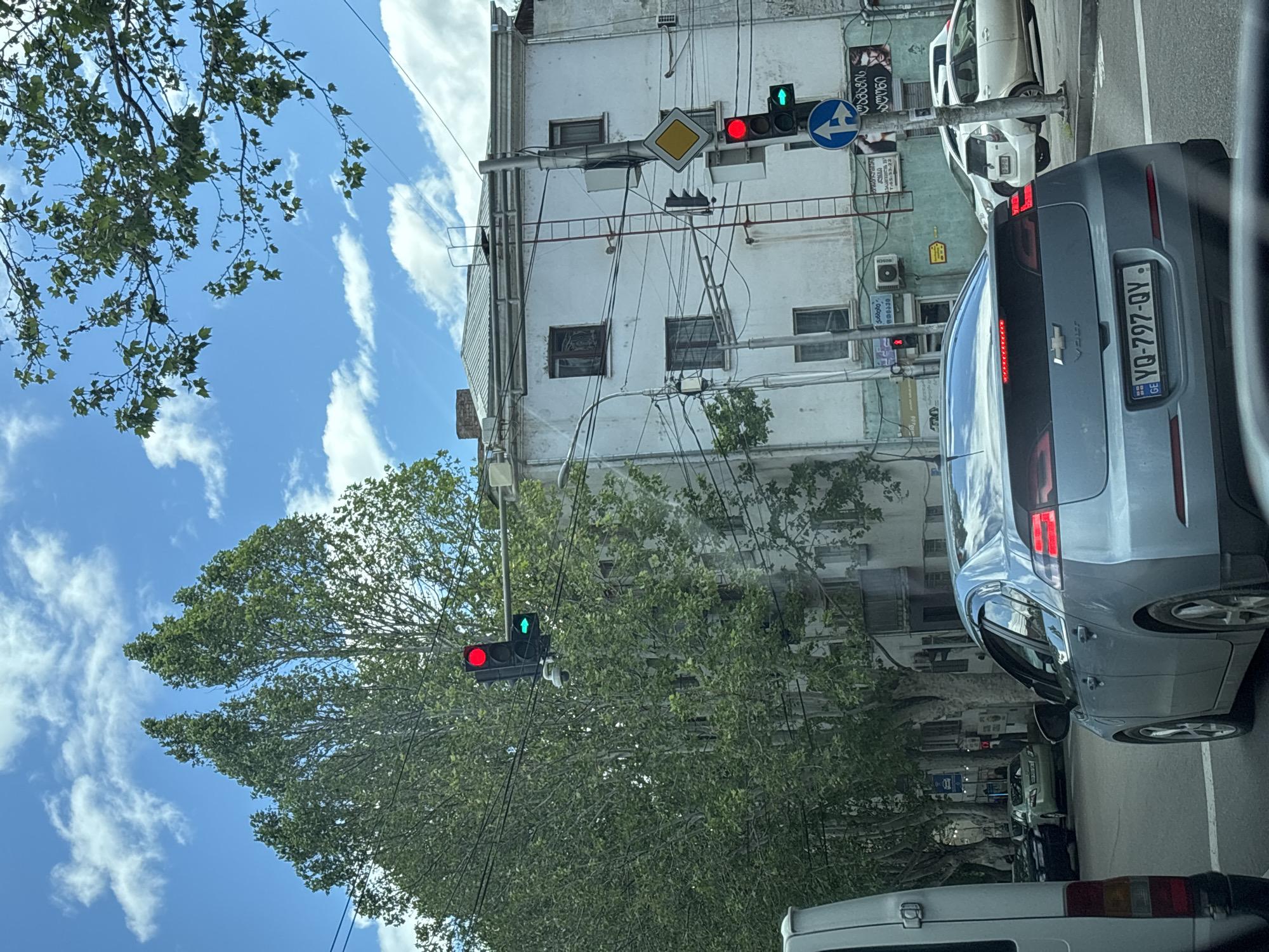 The image captures a street scene in Tbilisi, Georgia. A silver Chevrolet Volt is the primary subject, displaying a Georgian license plate (YQ-797-QY). Traffic lights are visible: one shows a red light, and another displays a green light for a left turn arrow. The street is lined with trees. Overhead wires crisscross the sky. An older white building stands adjacent to the street, with a yellow diamond warning sign and an arrow indicating a left turn lane. Other vehicles are parked along the street.