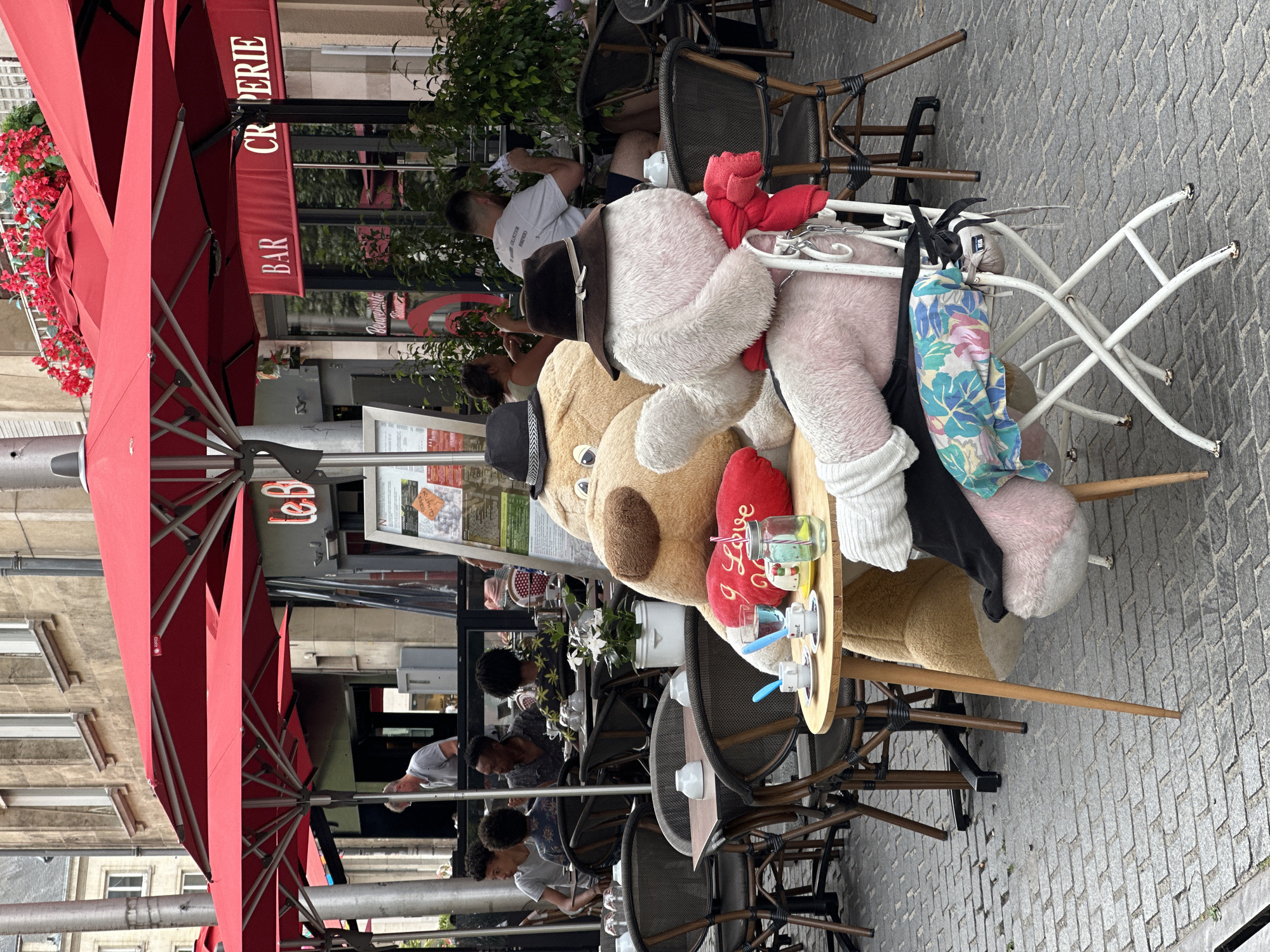 The image depicts an outdoor seating area of a cafe in Amiens, France. Two large plush teddy bears are seated at a small wooden table, facing each other. The bear on the left is tan and larger, while the bear on the right is pink and smaller. Both bears are wearing hats. The tan bear has a red heart pillow with "I Love" written on it. The table is set with two cups of coffee, a glass of water, and a straw. In the background, other patrons are seated at tables under red umbrellas. The cafe appears to be on a cobblestone street with buildings in the background. The overall scene suggests a playful or whimsical display at the cafe.