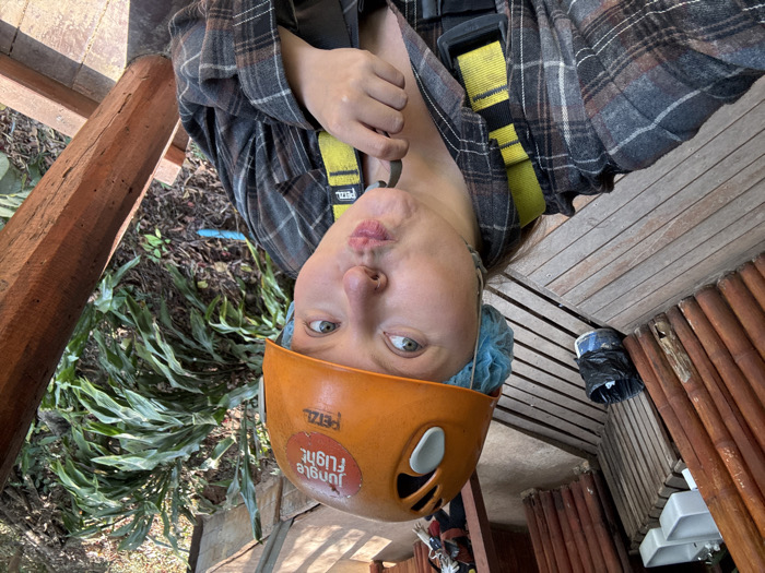 An eye-level portrait shows a person in an outdoor setting, positioned as if hanging upside down, though the image itself is rotated. The person, who appears to be an adult with fair skin and light-colored eyes, is looking directly at the camera with a playful expression, her lips pursed in a 'fish face'. She is wearing an orange safety helmet with white accents, underneath which a light blue, disposable hair net or cap is visible. A sticker on the helmet reads "JUNGLE FLIGHT" with a graphic of a bird. The brand "PETZL" is also visible on the helmet.
The person is wearing a grey and brown plaid long-sleeved shirt and a yellow safety harness, also bearing the "PETZL" brand name. The harness straps are visible across her shoulders and chest.
The background suggests an adventure activity in a jungle or forest environment. To the left (from the viewer's perspective, assuming the image is correctly oriented), there's lush green foliage with large leaves, indicative of tropical vegetation. A thick wooden beam, part of a structural element like a platform or tower, is prominent on this side. To the right, there's a wooden platform or structure, possibly part of a zipline station or observation deck, made of horizontal wooden planks. Further right and below, there are vertical wooden poles or slats, and some white sink basins are partially visible, suggesting a facility area. A black trash bag and a blue container are also seen on the wooden platform. The lighting suggests it is daytime.