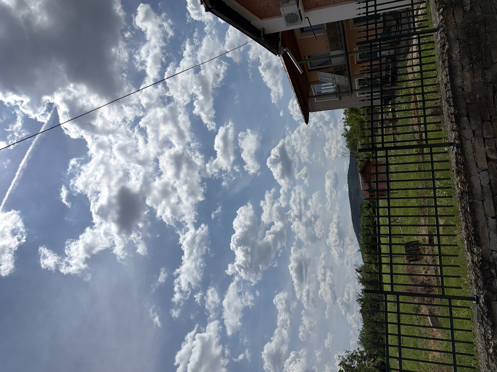 The image shows a view of a residential area with a house, a fence, and a landscape under a cloudy sky. The house is a multi-story building with an orange facade, a balcony, and an air conditioning unit on the exterior wall. A black metal fence runs across the foreground, separating the viewer from a grassy yard with some debris. In the background, there are trees, a smaller red-roofed building, and rolling hills. The sky is predominantly blue with a scattering of white and gray clouds. A thin black wire stretches diagonally across the upper portion of the image. The scene appears to be a sunny day. The location is identified as Bahovitsa, Bulgaria.