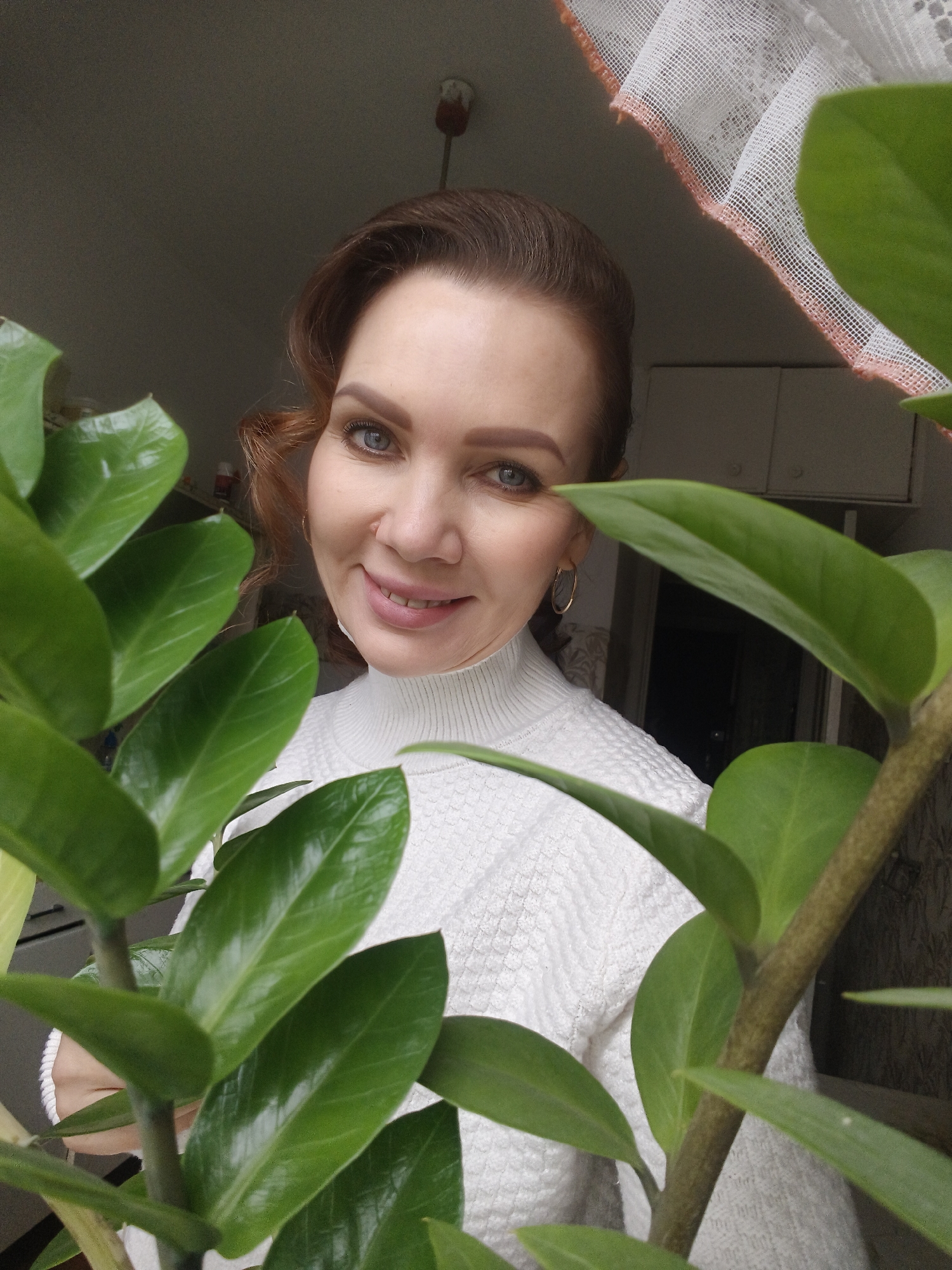 A woman with reddish-brown hair and blue eyes is posing indoors, partially obscured by a leafy green plant. She is wearing a white turtleneck sweater and gold hoop earrings, and she has a slight smile. The plant, with its thick, glossy leaves, dominates the foreground of the image. Behind the woman, a sheer white curtain hangs in front of a window. In the background, cabinets are visible. The location is Unknown, Unknown.