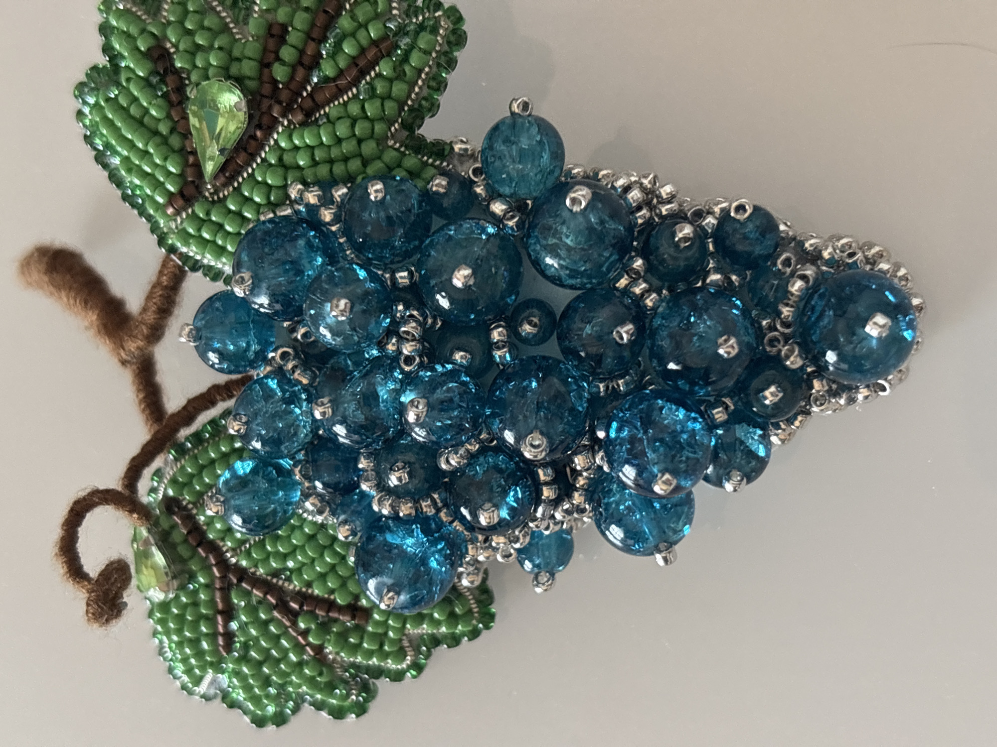 A close-up, top-down shot shows a beaded grape cluster brooch. The brooch features several large, translucent, faceted blue beads representing grapes, surrounded by smaller silver beads. Two green beaded grape leaves with brown beaded veins and a brown fuzzy stem complete the design. A light green faceted jewel is attached to each leaf. The brooch rests on a white, reflective surface. There are no people or indications of location in a city.