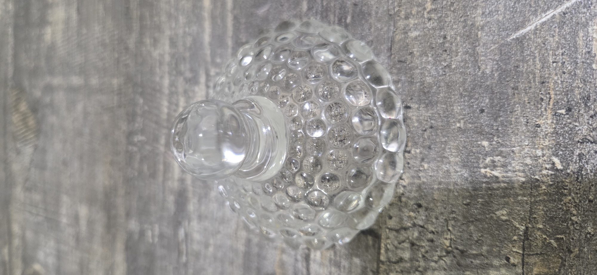 A clear glass lid with a bubble pattern and a spherical knob sits on a dark, textured wooden surface. The lid has a rounded, dome-like shape with a scalloped edge. The bubble pattern is raised and covers the entire surface of the lid, with each bubble appearing to be a small, rounded indentation. The knob is a simple, clear glass sphere attached to the center of the lid. The wooden surface has a distressed or weathered appearance, with visible grain and variations in color, appearing as shades of gray and brown. The lighting suggests an overhead light source, casting soft shadows and highlighting the transparency and texture of the glass. There are no people, animals, or other objects visible in the image. The scene is static and focused solely on the lid and its immediate surroundings. The location in Lipetsk, Russia, is not discernible from the visual information provided.