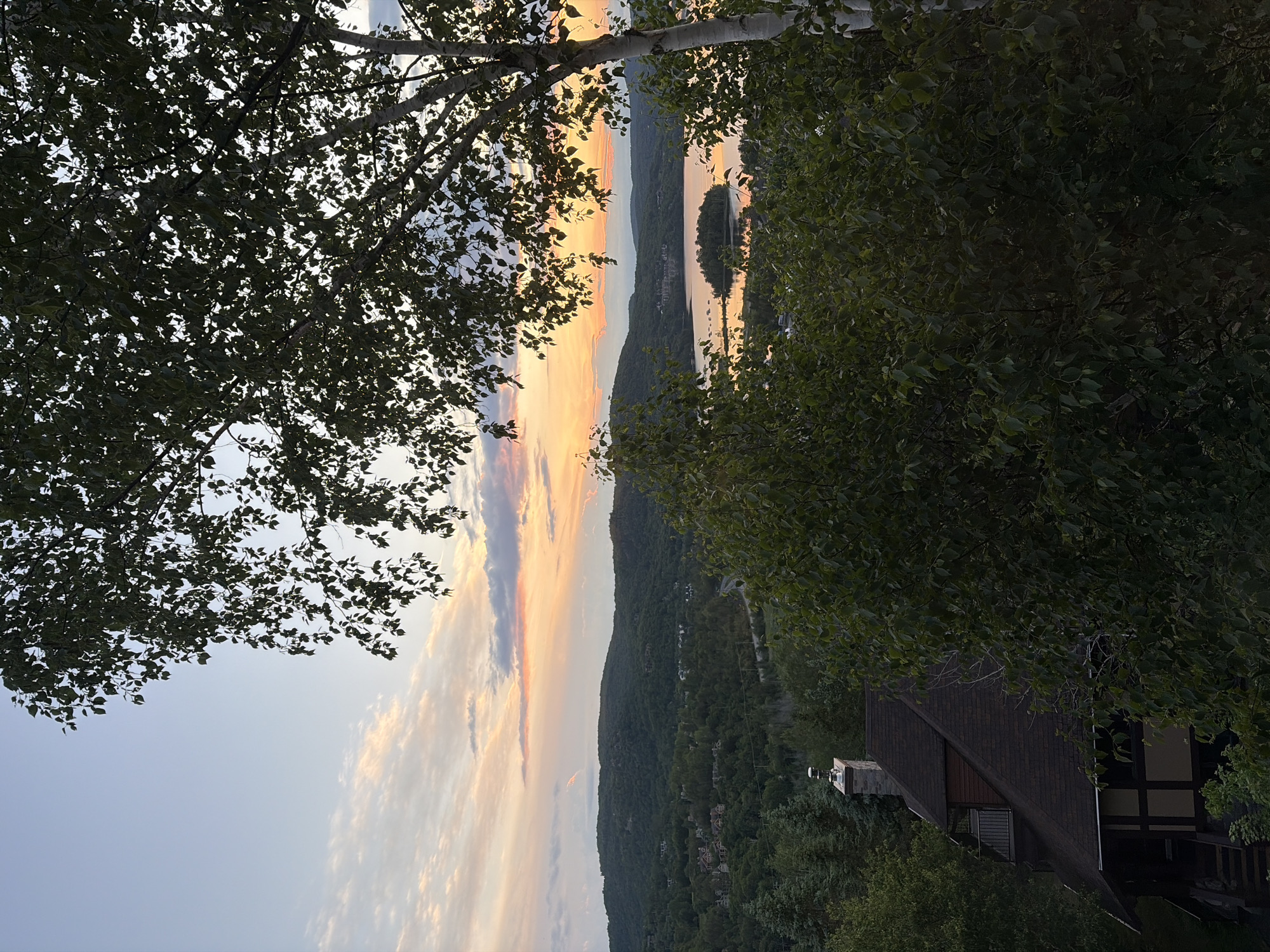 The image depicts a serene landscape in Mont-Tremblant, Canada, during a sunset. The foreground is dominated by lush green trees, with a portion of a dark-roofed house with a stone chimney visible on the left. The middle ground features rolling green hills dotted with houses, leading to a tranquil body of water reflecting the warm hues of the sky. The background showcases a majestic mountain range under a sky painted with a gradient of soft blues, oranges, and pinks from the setting sun, with scattered clouds adding texture. There are no people or animals visible in the scene. The overall impression is one of peaceful natural beauty and a suburban dwelling nestled within the landscape.