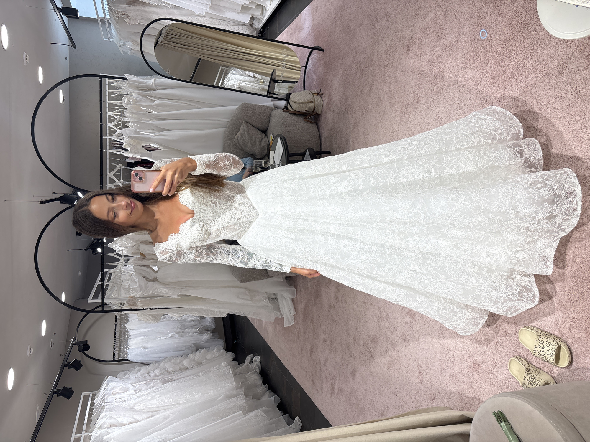 A woman wearing a white lace wedding dress is taking a selfie in a bridal shop. The shop features racks of dresses, mirrors, and a pink carpet. The location is not specified, and there is no indication that it is Saint Petersburg, Russia.