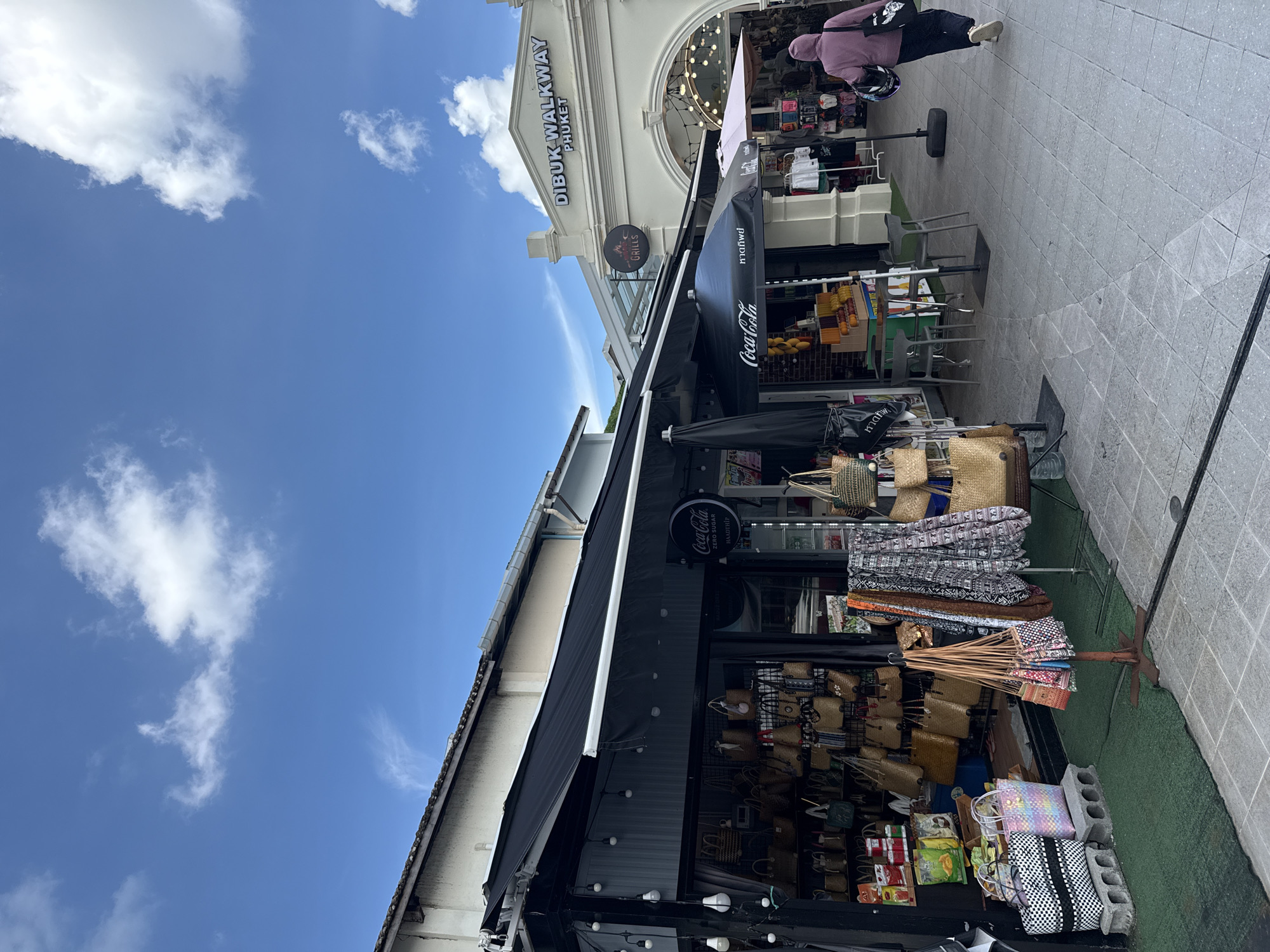 This is a medium shot image of Dibuk Walkway in Phuket City Municipality, Thailand, on a partly cloudy day. The walkway is paved with grey tiles, and there are several market stalls lining the path. One stall in the foreground is selling an assortment of woven bags and clothing. The stall has a black awning, and string lights are hung along its edge. Next to this stall, an outdoor seating area with two grey tables and chairs is set up under a black Coca-Cola umbrella. Further down the walkway, a building with an arched entrance is visible, with a sign that reads "DIBUK WALKWAY PHUKET". A person wearing a purple hooded jacket and dark pants is walking away from the camera down the walkway. They are carrying a black bag over their shoulder.