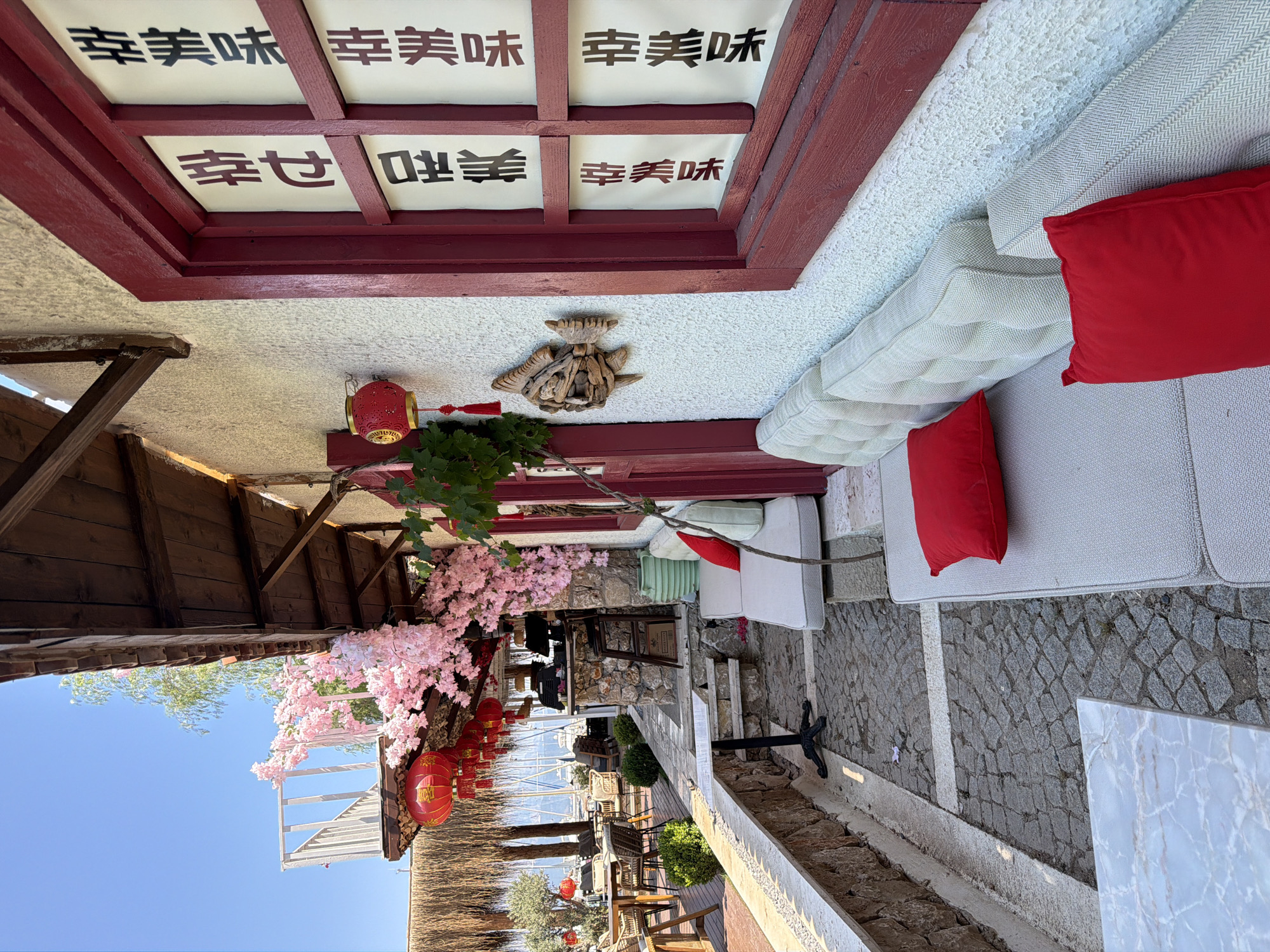 The image shows an outdoor seating area of a restaurant in Kemer, Turkey, decorated with a Japanese theme. The seating consists of plush, light-colored sofas with red accent pillows. The building features a white stucco exterior with dark red trim around the windows, which display white panels with black Chinese characters. Decorative pink cherry blossoms hang from an overhang, and red lanterns are visible, adding to the Asian aesthetic. A carved wooden fish is mounted on the wall. The ground is paved with cobblestones and white stone borders. In the background, tables and chairs are set up under a thatched roof, and a view of the sea with sailboats can be seen. The sky is clear and blue. There are no people visible in the image.