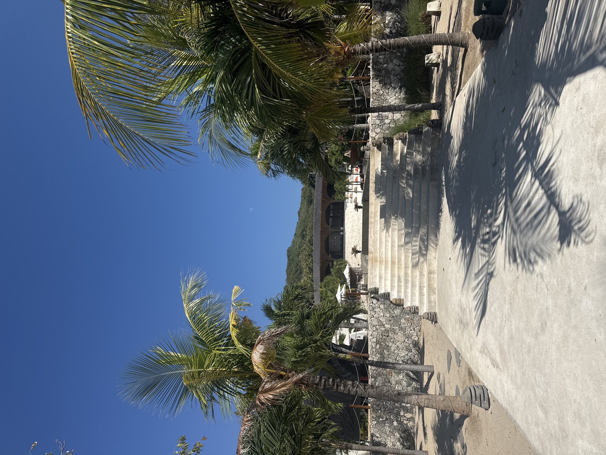 A wide-angle, eye-level photograph captures the entrance to a tropical resort or facility on Nusa Penida, Indonesia, under a clear, bright blue sky. A faint crescent moon is visible in the upper-center sky. The scene is dominated by a central concrete pathway leading to a broad, ascending concrete staircase. Numerous palm trees with green fronds, some exhibiting slight browning, frame the pathway and staircase, extending from the foreground into the midground. Distinct, dark shadows of palm fronds are cast across the light grey concrete pathway in the foreground, particularly on the right side. The ground flanking the pathway consists of light-colored sand or fine gravel, interspersed with sparse green vegetation. Stacked stone decorative elements, resembling small totems or planters, are positioned along the pathway edges and at the base of some palm trees.
The multi-tiered concrete staircase, symmetrical to the central axis, ascends towards a large, open-air building in the background. Along the base of the staircase and flanking it, a low wall constructed from irregular grey and tan stones separates the lower pathway level from the elevated area. A simple rope railing is visible atop sections of this stone wall. The elevated area, accessed via the stairs, features more palm trees and several white, open sun umbrellas, suggesting outdoor seating or lounge spaces.
The main building at the top of the staircase has a dark, sloped roof and features prominent dark wooden arches or beams that define its open facade. Its interior appears shaded. Behind and above this structure, a lush, green, forested hill or small mountain rises under the blue sky. No people are visible within the frame.
