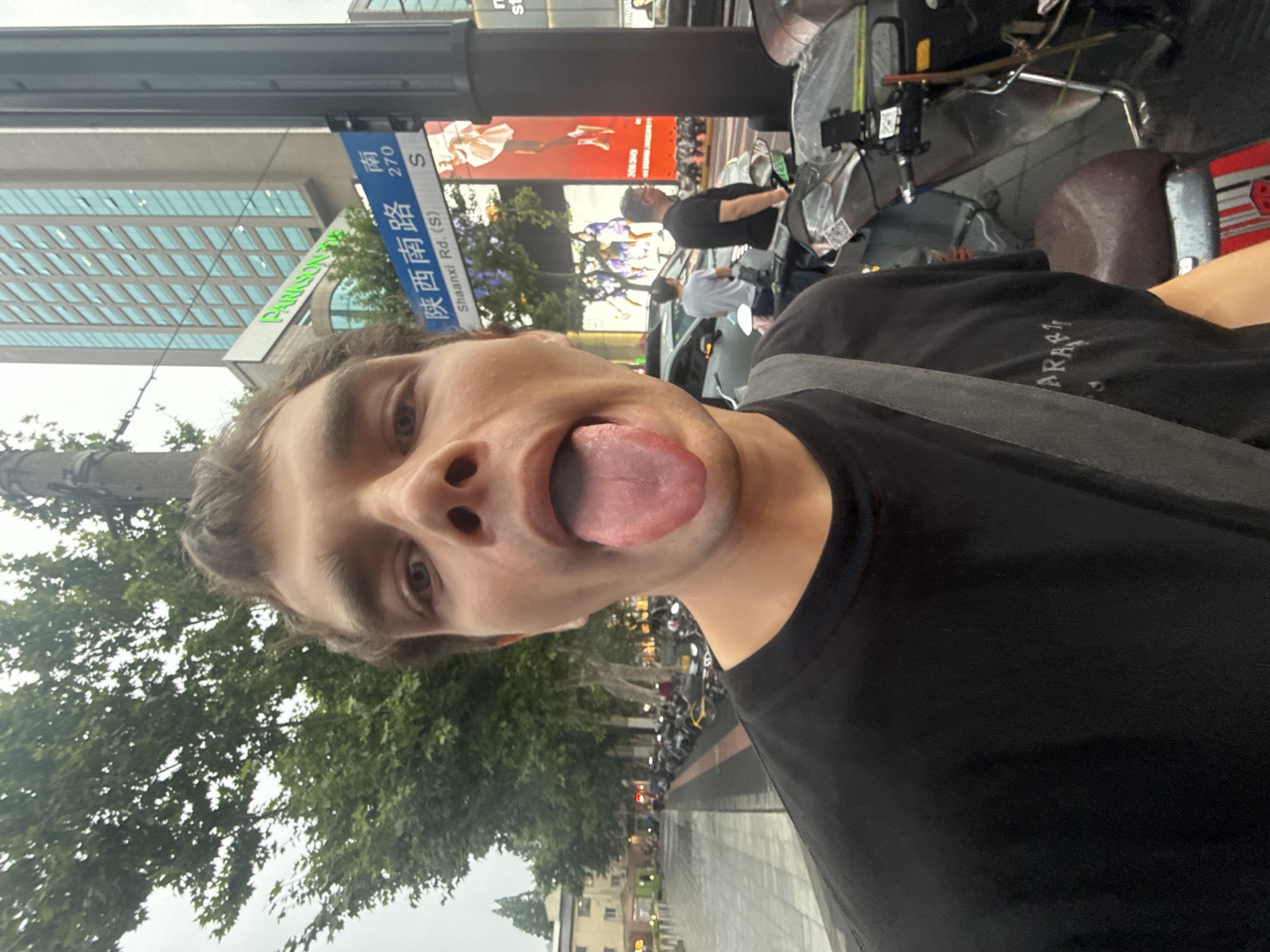 A man is featured in a close-up selfie, making a playful expression with his tongue sticking out. He has light skin and brown hair, and is wearing a black t-shirt with the partial text "ARAS" visible on it. The photo is taken on a city street during what appears to be an overcast day.
In the background, a blue street sign clearly identifies the location as "陕西南路" (Shaanxi Nan Lu), also labeled "Shaanxi Rd. (S) 270 S", confirming it as Shaanxi South Road in Xuhui District, China. A modern building with reflective glass windows stands prominently behind the street sign, and a green sign with "PARISON" (likely part of "PARKSON") is also visible.
The street is lined with green trees. Other people can be seen in the middle ground, alongside parked motorcycles or scooters and a partial view of a car. A large red billboard is also visible further down the street. The overall setting is a bustling urban environment.