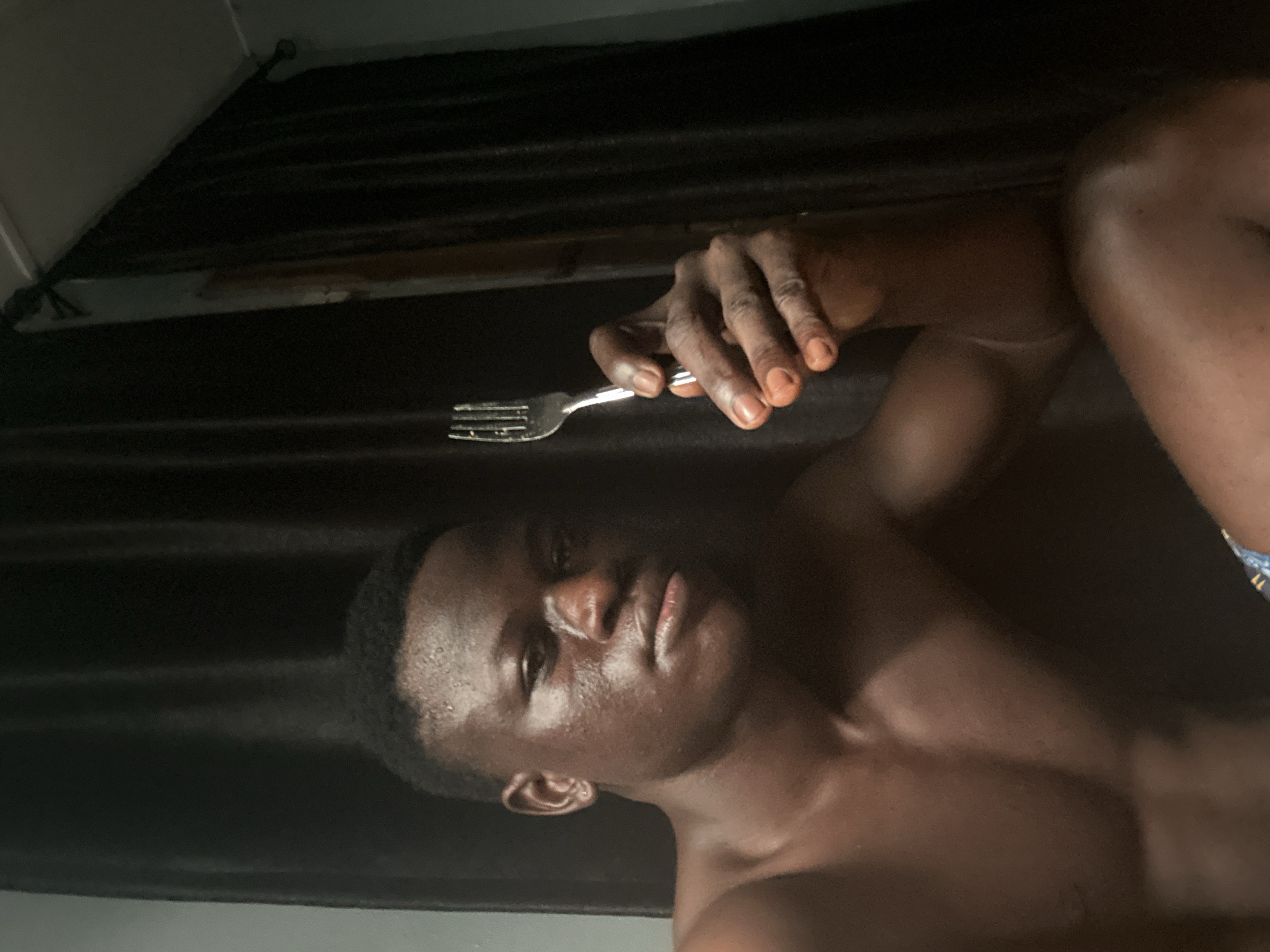 The image features a young Black man lying down in a dimly lit room, with a dark curtain as the backdrop. He is holding a silver fork in his right hand, positioned above his head. His skin appears slightly damp, suggesting perspiration. The man's face is visible, turned towards the camera, with his eyes closed and lips slightly parted. His body is partially visible, showing his chest and shoulder. The overall mood is intimate and personal, possibly captured during a moment of relaxation or rest. The limited lighting creates a dramatic effect, highlighting his features and the texture of his skin. There are no visible text elements in the image. The location context of Badagry, Nigeria, is noted, but there are no specific visual cues in the image to confirm this location.