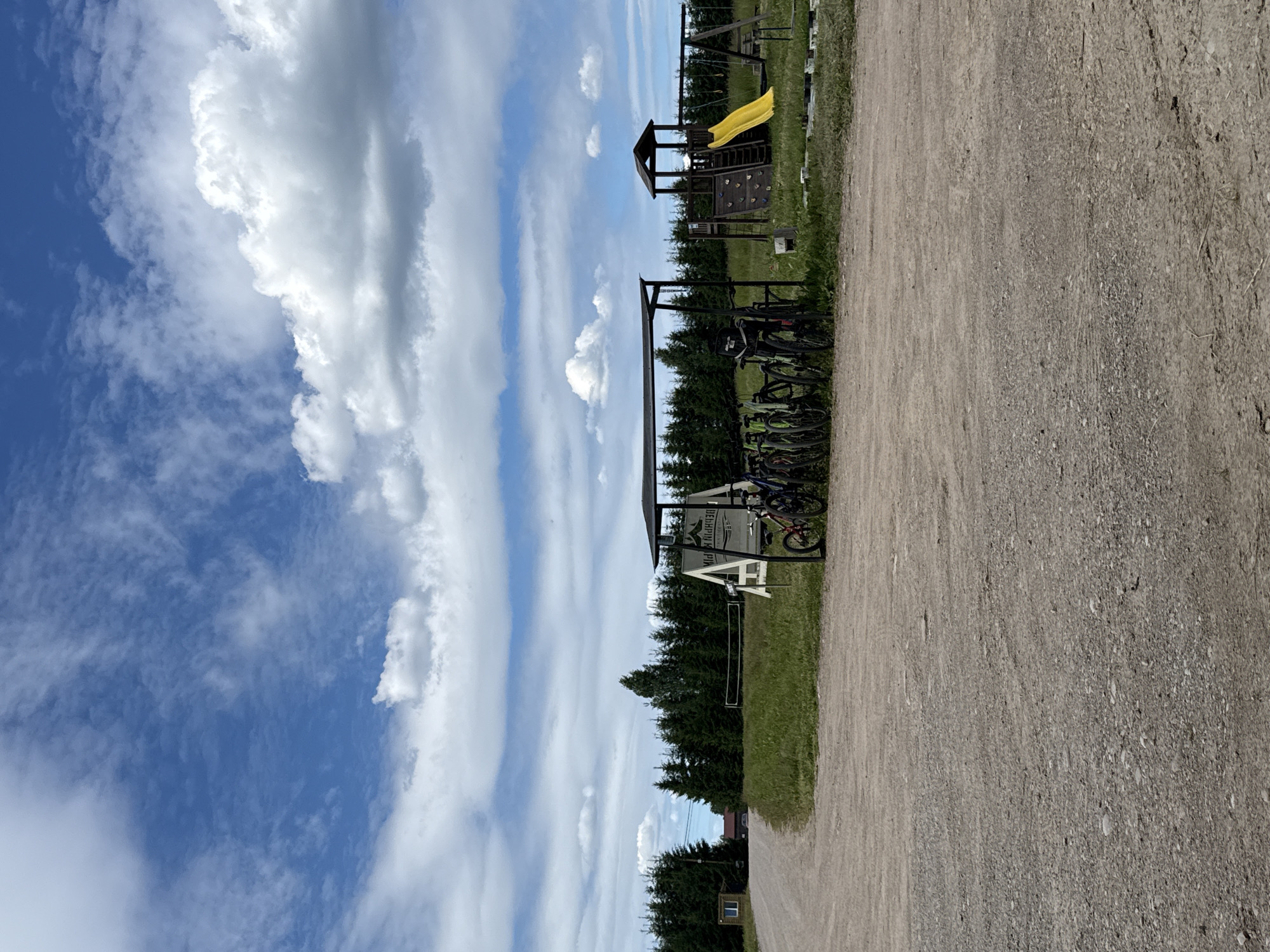This image shows a rural scene in the city of Zamosh'e (Zaklinskoe S/P), Russia. The foreground is a gravel road leading to a grassy area with a bicycle rack under a dark shelter. Several bicycles are parked in the rack. Behind the bicycles, a playground structure with a yellow slide, swings, and climbing wall is visible. A volleyball net stands further back in the grassy field, bordered by a dense line of dark green coniferous trees. In the distance, a small building with a window can be seen to the left of the trees. The sky above is blue with scattered white clouds.
