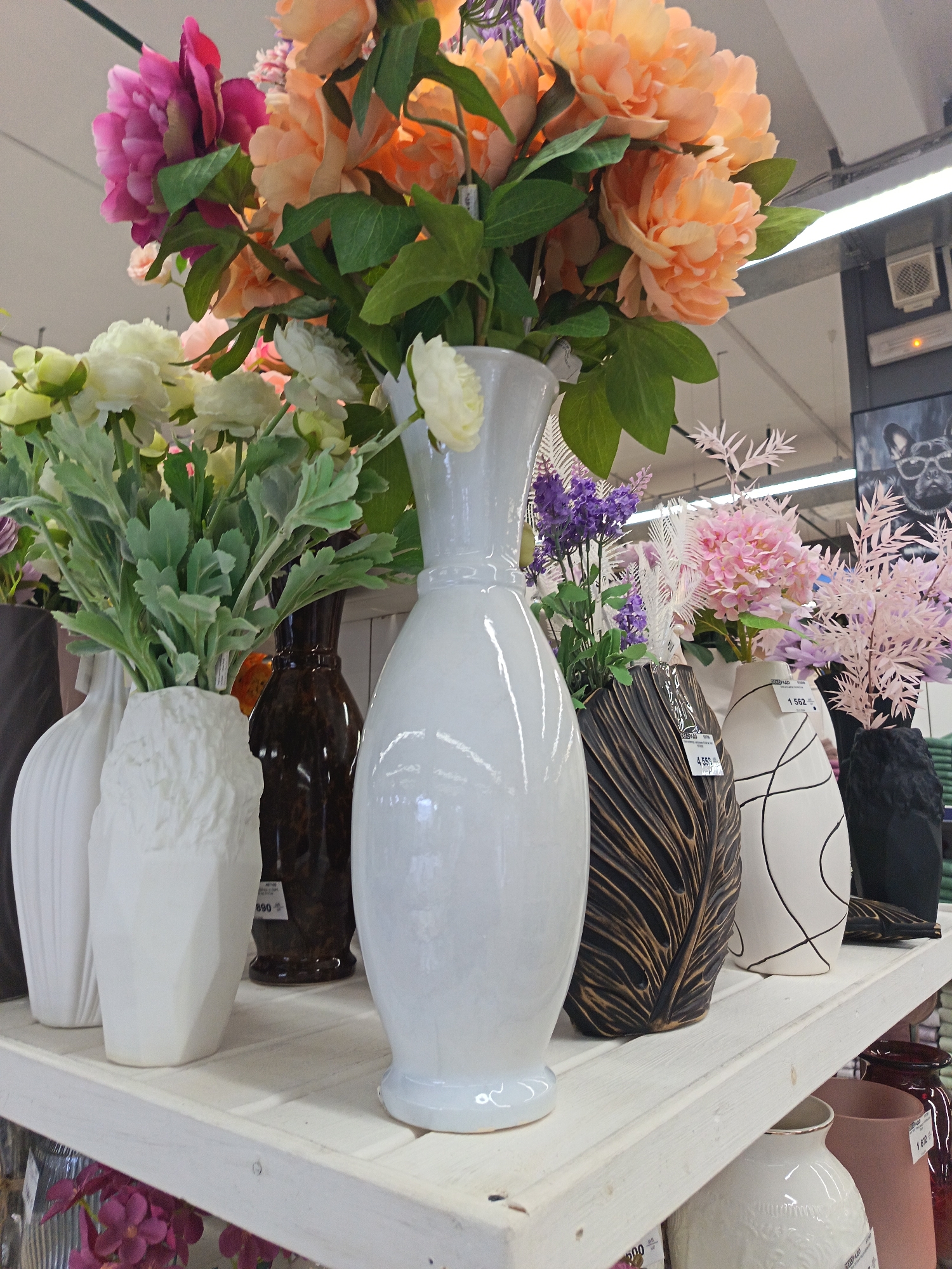 This photo shows a shelf of vases with flowers. There are at least 10 vases of different shapes and sizes. Some of the vases are white, some are brown, and some are black. The flowers in the vases are of different colors, including pink, orange, purple, and white. The shelf is made of wood and is painted white. The photo appears to have been taken in a store, as there are price tags on some of the vases. There is a sign visible in the background, with text in Russian (based on the file name, "Nizhnevartovsk, Russia"), which suggests the photo was taken in a store in Nizhnevartovsk, Russia.