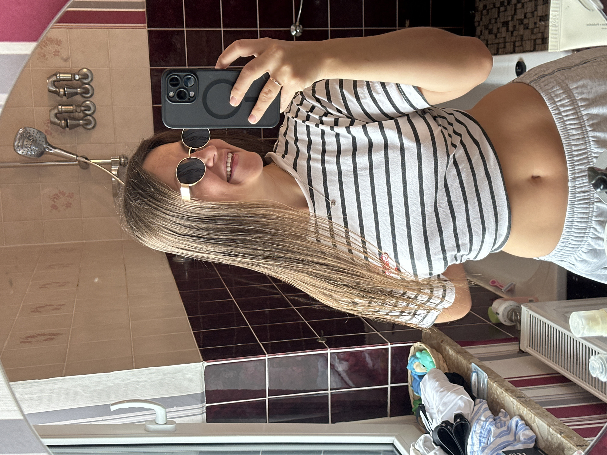 A young woman is captured in a mirror selfie taken in a bathroom, the image itself rotated 90 degrees counter-clockwise. She is smiling broadly, revealing white teeth, and wearing round, gold-rimmed sunglasses. Her long, light brown hair cascades over her shoulder. She is dressed in a white and black striped crop top, revealing her midriff, and light grey athletic shorts with an elastic waistband.
The woman holds a dark-colored smartphone, likely an iPhone given the multi-lens camera system and visible MagSafe ring, in her right hand, positioned to take the photo. A simple silver ring is visible on her ring finger.
The bathroom features tiled walls, with an upper section of light beige or pinkish tiles and a lower section of dark brown or maroon tiles separated by a patterned border. A metallic shower head and two ornate metallic faucet handles are visible on the upper tiled wall. Below, a white window frame with a handle is seen, and a white radiator is partially visible. Various toiletries and bottles are arranged on a counter or shelf next to a white sink. A basket or bin filled with clothes, including striped blue and white items and black shoes, sits near the window. A pink razor and other small items are also discernible on the surfaces. The setting suggests an indoor space, likely a residence in Fredenbeck, Germany, during daylight hours, as indicated by the natural light. No discernible text is visible in the image.