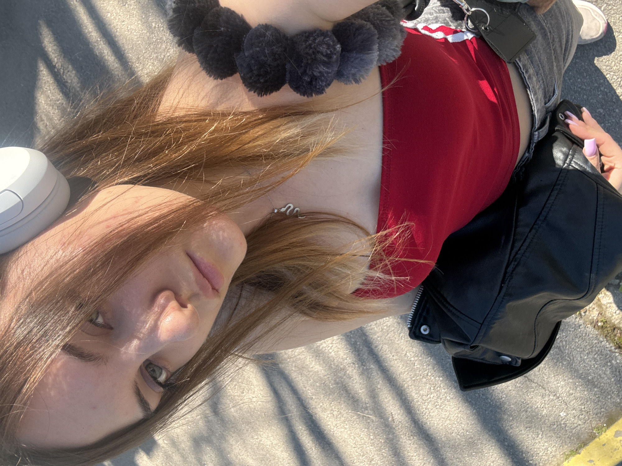 A woman is lying on a paved surface, likely a sidewalk, on a sunny day. Her head is oriented towards the top-left of the frame, and her body extends diagonally across the image. She has light brown hair, green eyes, and fair skin. She is wearing a red top, gray jeans, and a black leather jacket is near her. A necklace is visible on her neck. A black, pom-pom-like accessory is around her neck. A white, oval-shaped object is next to her head. The location appears to be outdoors, with shadows visible on the pavement. There is no definitive visual information in the image to place the location specifically within the city of Sofia, Bulgaria.