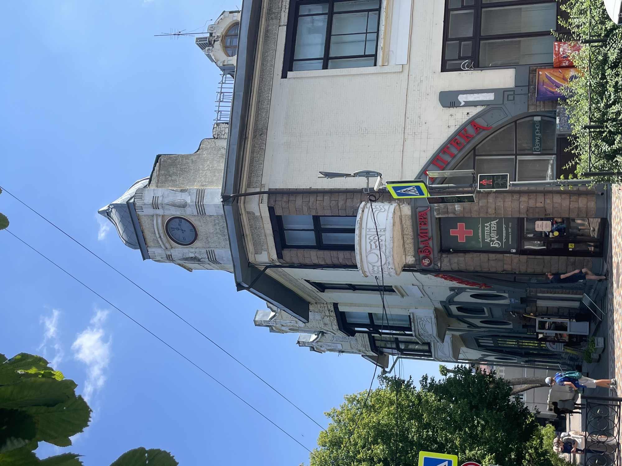 The image shows a street view in the city of Stavropol, Russia, focusing on a multi-story building with distinctive architectural features. The building is cream-colored with dark-framed windows and decorative stone accents. A prominent clock is visible on a turreted section of the building. A pharmacy, identified by a red cross symbol and Cyrillic text, is located on the ground floor, with a street signal showing a red pedestrian light. People are walking on the sidewalk, and trees line the street, suggesting a daytime scene. Overhead power lines are also visible against a clear blue sky with scattered clouds. The overall impression is of an urban environment on a sunny day, highlighting historical architecture and daily life.
