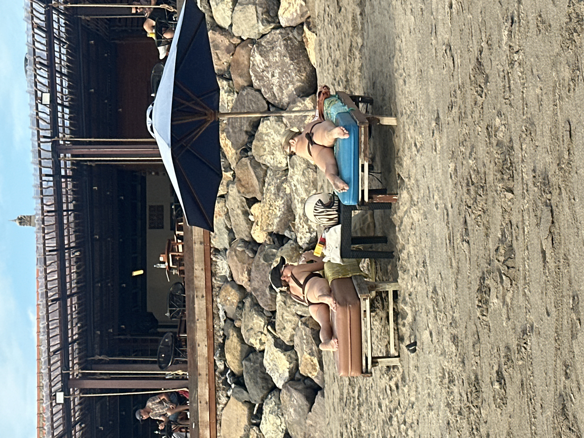 Two women are lying on lounge chairs on a sandy beach in front of a rocky embankment. Behind the embankment is a wooden structure with a thatched roof, likely a restaurant or bar. A large blue umbrella shades one of the lounge chairs. The woman on the left wears a black bikini and a baseball cap, while the woman on the right wears a brown bikini. A small table sits between the two chairs with a helmet and a bag on it. The scene appears to be in North Kuta, Indonesia.