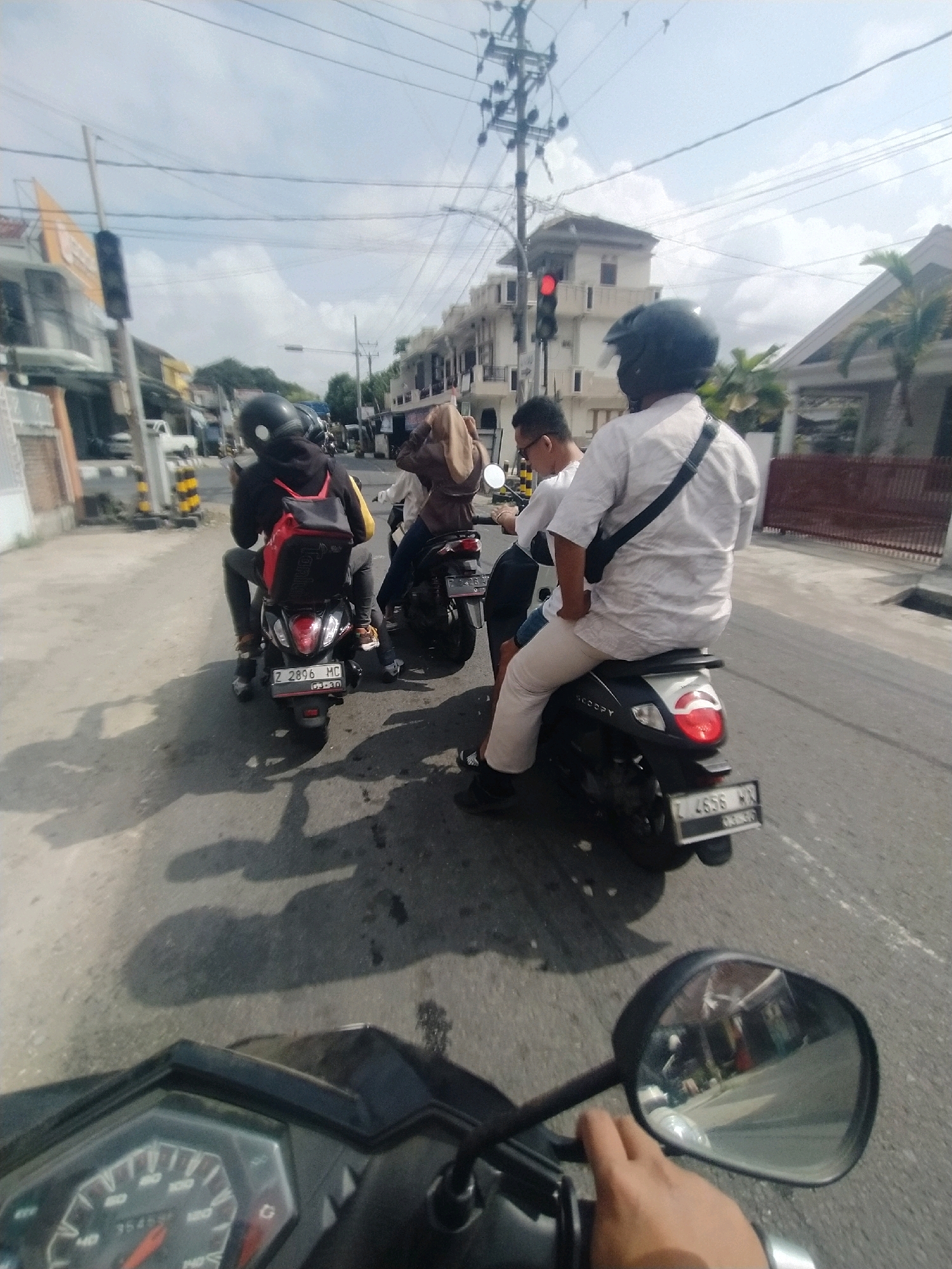 The image captures a street scene in Desa Karangbenda, Indonesia, with multiple motorcycles in motion. In the foreground, a hand is visible on the handlebars of a motorcycle, with a speedometer display in the lower left corner. The perspective suggests the photo is taken from the viewpoint of someone riding this motorcycle.
Behind the foreground motorcycle, two other motorcycles are visible. The first, slightly to the left, carries two individuals wearing helmets. The rider in front wears a black helmet and a black hoodie with a red backpack. The passenger behind is obscured by the rider. This motorcycle has a license plate reading "Z 2896 MC".
Further ahead and to the right, another motorcycle is seen with two occupants. The rider wears a white shirt and a black shoulder bag, with a dark helmet. The passenger wears a light brown headscarf and a light brown jacket. This motorcycle has a license plate reading "L 4656 WH".
The environment appears to be an urban or suburban street with buildings lining both sides. Power lines are strung across the sky. A traffic light is visible in the mid-ground, displaying a red light. The road surface is asphalt, with shadows of the motorcycles and their riders cast upon it. The sky is partly cloudy.