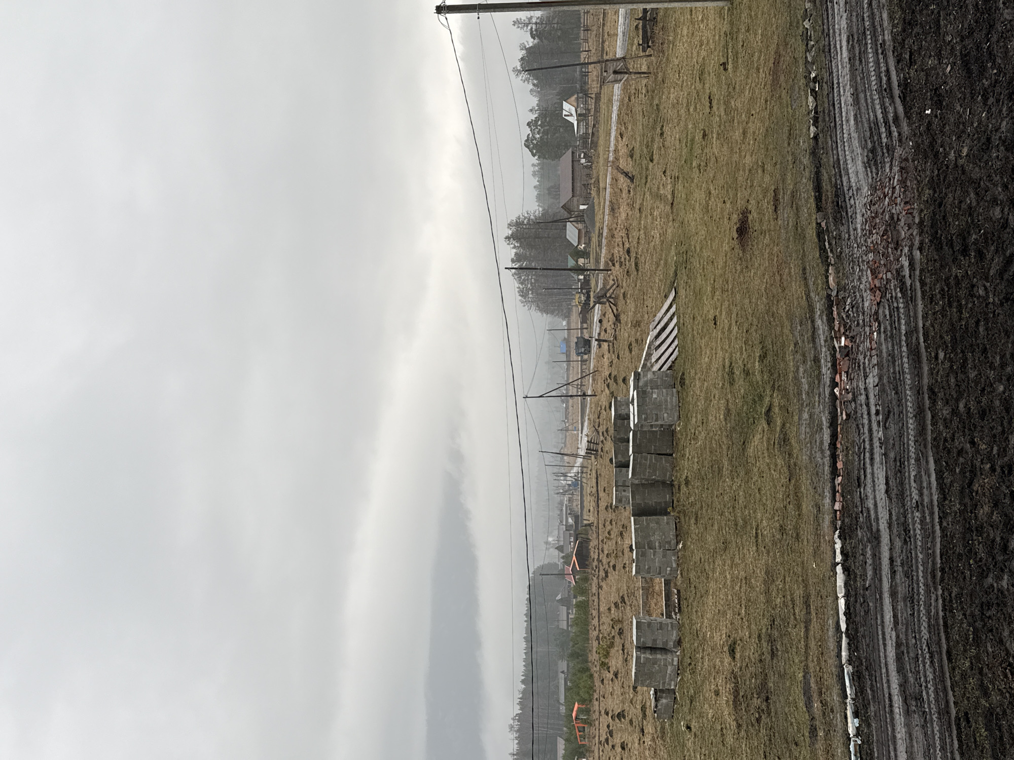 The image shows a rural scene in Tyulyuk, Russia, under an overcast sky. In the foreground, there is a muddy track with visible tire marks, bordered by a grassy area with scattered bricks. A stack of rectangular concrete blocks lines the edge of the grass. Beyond the blocks, there is a black and white striped barrier. In the distance, a scattering of houses and trees is visible, fading into the misty atmosphere. Power lines stretch across the sky.