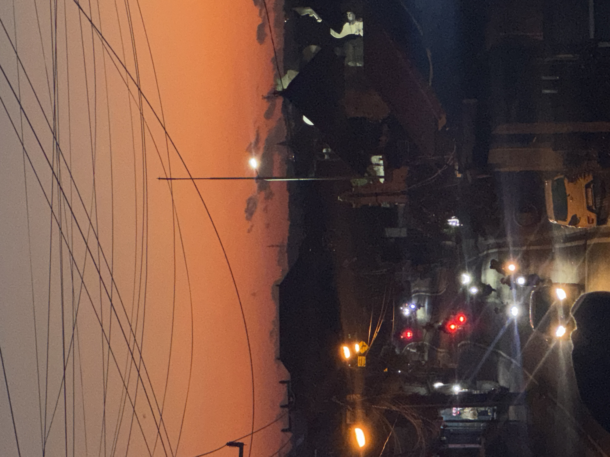 This image depicts an urban street scene in Kuta Selatan, Indonesia, during the transition from sunset to twilight. The upper third of the frame is dominated by a clear, vibrant orange-red gradient sky, against which numerous dark utility wires crisscross irregularly. A single tall, slender utility pole stands prominently on the right side, extending from the midground into the upper sky.
The scene is viewed from an elevated perspective, looking down onto a main thoroughfare. The midground features a busy street with active vehicle traffic. Multiple vehicles are visible: numerous motorcycles, identified by single bright white or yellow headlights and red taillights, are distributed along the road, some appearing to be in motion. At least two cars are present; one white car with bright, illuminated headlights faces the viewer in the lower-center, and a yellow hatchback is parked on the right side of the road.
Artificial lighting is prevalent. Several orange streetlights illuminate the immediate vicinity of the road, and additional white lights are visible on utility poles. Buildings line both sides of the street, appearing as dark silhouettes with some illuminated interior spaces or signs casting light. Cluttered electrical wiring is dense above the street level, connecting to poles and structures.
In the background, beyond the immediate street and buildings, dark silhouettes of more distant structures and subtle terrain features are visible against the fading orange light of the horizon. Faint, dark clouds are scattered across the upper sky. The overall scene is characterized by active urban evening life with significant artificial illumination against a colorful dusk sky.
