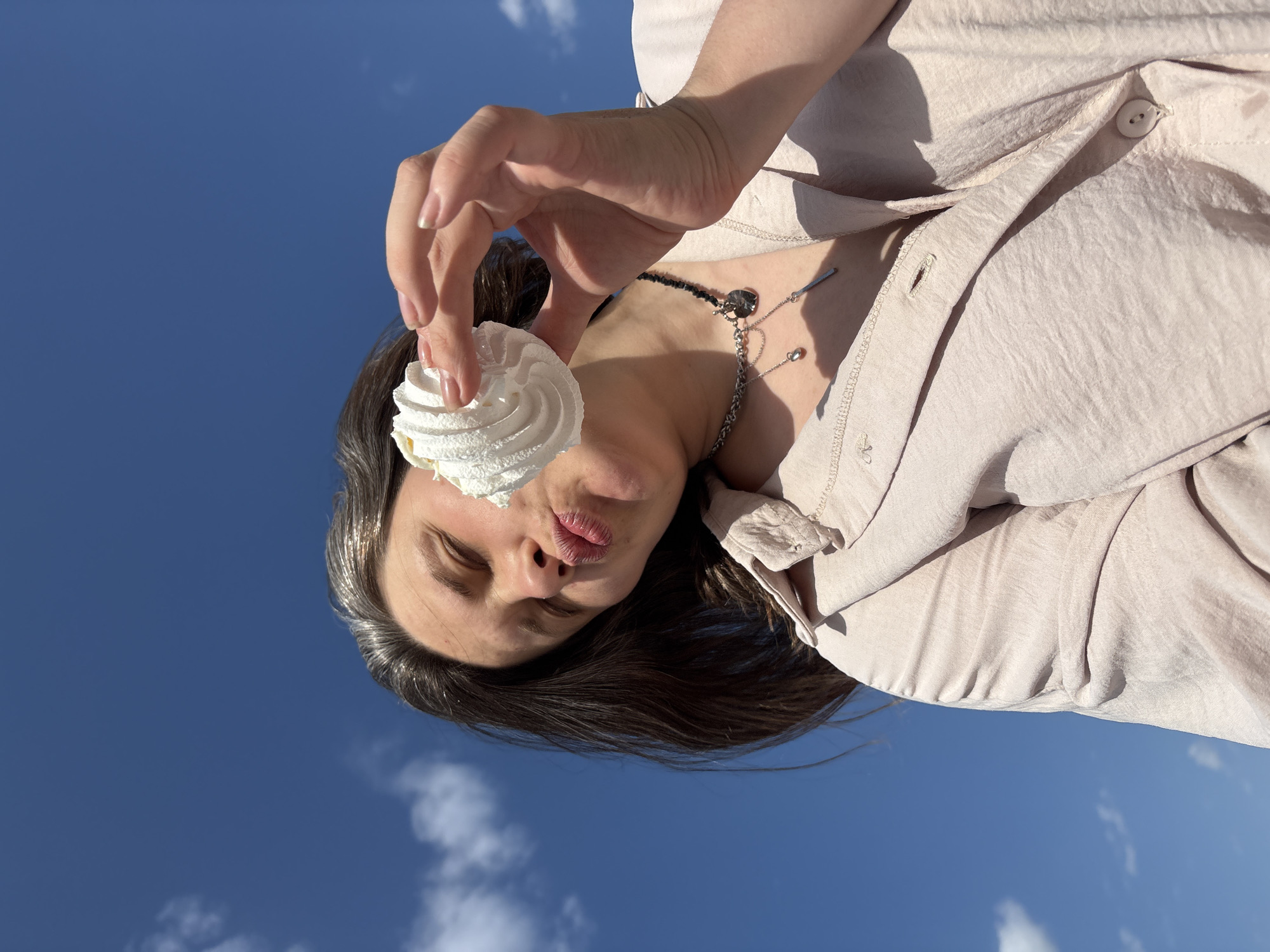 A rotated, low-angle, eye-level shot shows a person holding a white meringue cookie in front of their face. The background is a bright blue sky with a few scattered white clouds. The person appears to be female with dark hair, fair skin, and is wearing a light pink button-up shirt and a two-strand necklace. The location is indeterminate, no information available indicates this picture was taken in Chaikovsky, Russia.