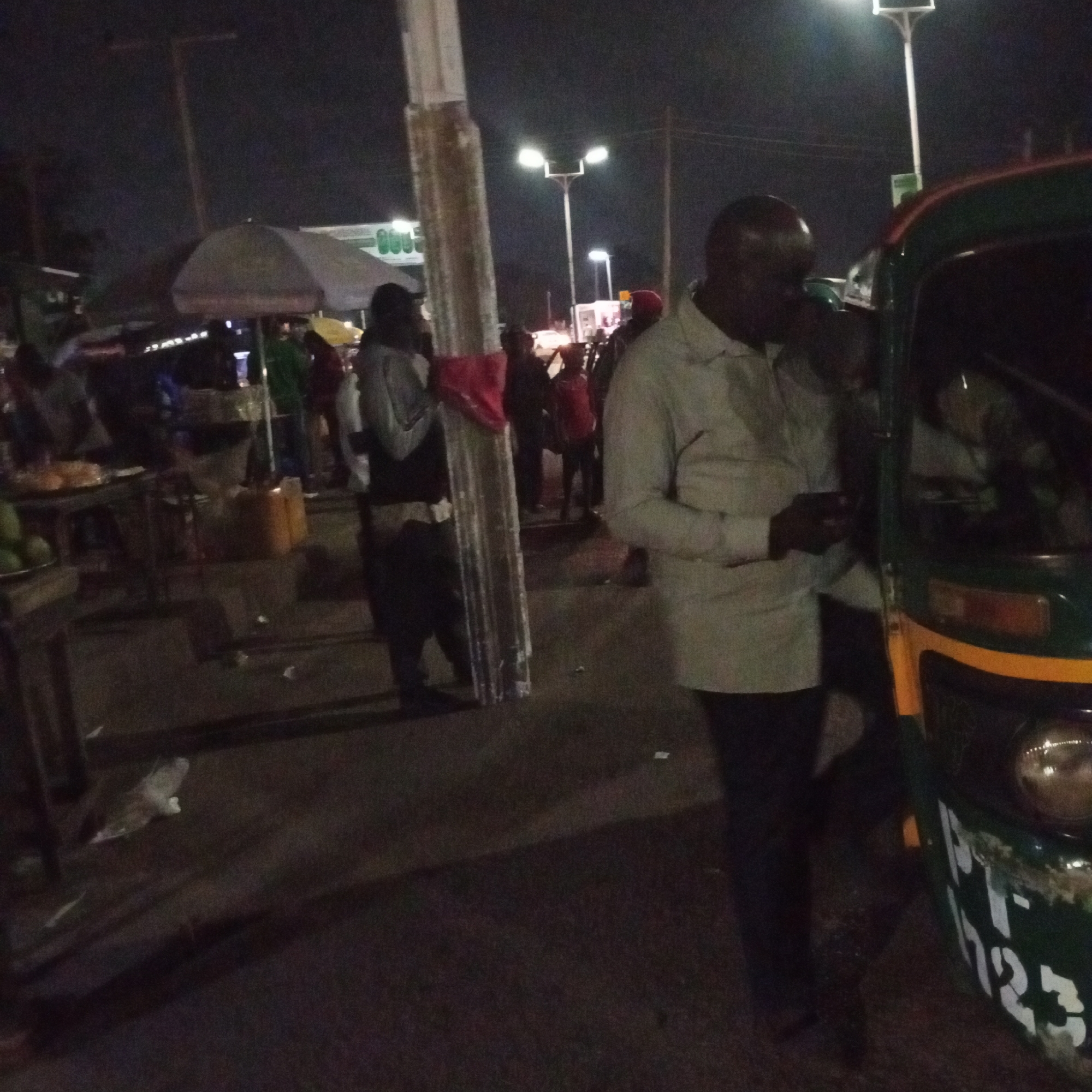 This is a nighttime street scene in Bukuru, Nigeria, likely a market area. A man in a light-colored shirt and dark pants stands next to the front of a green and yellow auto-rickshaw, holding a dark object in his right hand. The auto-rickshaw has a license plate with white numbers and letters that appear to read "PT-T 723". In the background, other individuals are visible, some standing near stalls or other vehicles. There is a large, light-colored umbrella or canopy to the left, and streetlights illuminate the scene. The ground is paved and appears somewhat uneven, with scattered debris. To the far left, a table with produce, possibly mangoes, is visible.