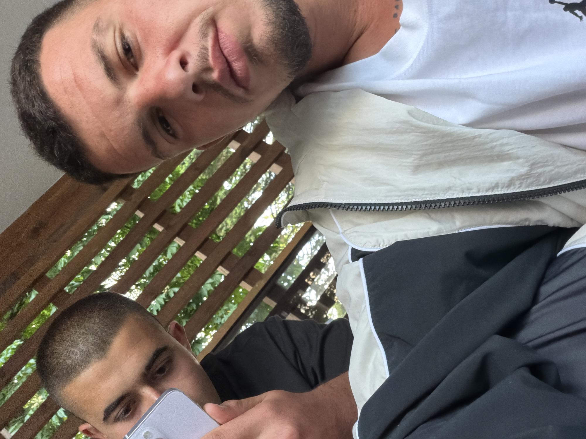 The image is a tilted, close-up shot featuring two men in an outdoor or semi-outdoor setting in Razlog, Bulgaria, likely taken as a selfie.
The man in the foreground, positioned at the top of the frame, has short dark hair and a light beard. He looks directly at the camera with a relaxed, subtle smile. He wears a white t-shirt with a visible black "Jumpman" logo on the shoulder, layered under a cream or off-white jacket with black panels and a zipper running down the front.
Below and slightly behind him, at the bottom of the frame, is another man with a very short, buzzed dark haircut. He is engrossed in a light-colored smartphone he holds, with its camera module clearly visible on the back. He wears a dark-colored (likely black) t-shirt.
The background shows a slatted wooden structure, possibly a pergola or trellis, with bright green foliage and patches of clear sky visible through the gaps. The bright lighting indicates it is daytime and likely sunny. The overall scene suggests a casual, outdoor environment.