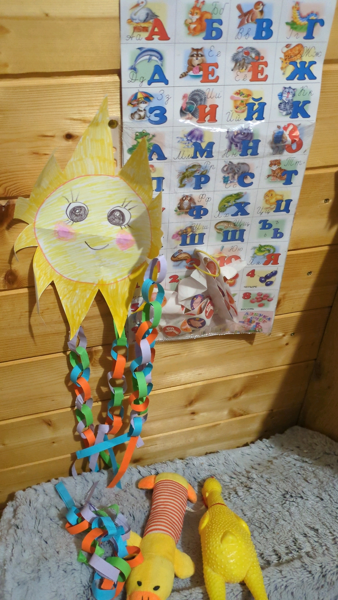 The image shows a wall covered with wooden planks, possibly in a child's room or playroom. A large, handmade paper sun with a smiling face hangs on the wall, with colorful paper chains dangling from it. To the right of the sun is a poster with Cyrillic letters and animal illustrations, likely an alphabet chart. Below the sun and to the left, more paper chains are scattered on a gray, fuzzy surface, which appears to be a rug or blanket. Two yellow rubber duck toys are also visible on the rug, one with red and white stripes. The lighting suggests an indoor setting, possibly with natural light coming from the side. There is no visible indication of the location being in Chișinău, Moldova, or any other specific city. The scene is static and depicts decorative elements and toys.