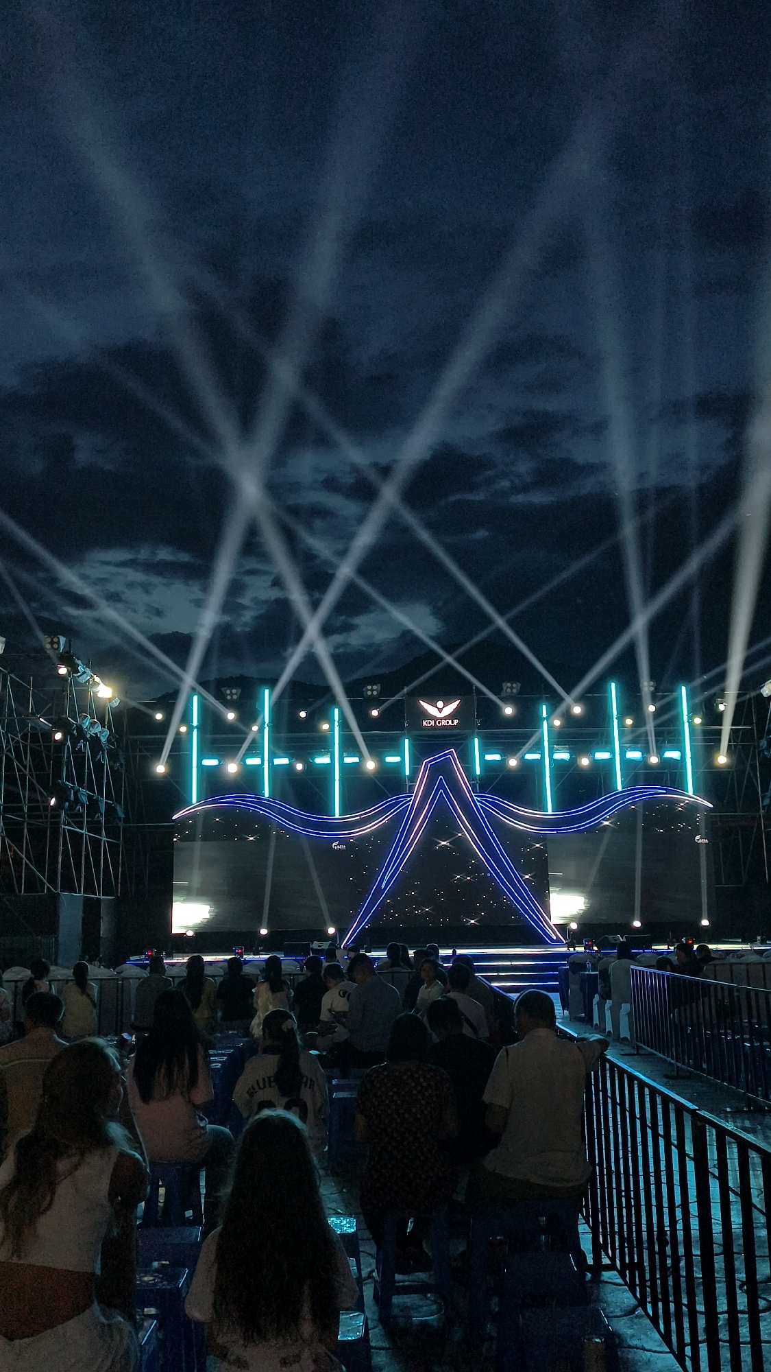 The image depicts an outdoor stage setup at night, likely for an event in Nha Trang, Vietnam. A large stage is illuminated by numerous bright white spotlights that fan out into the dark, cloudy sky. The stage itself features a prominent LED screen backdrop and is framed by blue and white neon lighting, forming a stylized star or wing-like shape. The logo "KDI GROUP" is visible above the stage. In the foreground, a crowd of people, mostly seen from behind, are seated on small blue chairs facing the stage. Some individuals are standing near a metal barrier on the right side of the frame. The overall atmosphere suggests a public performance or gathering.