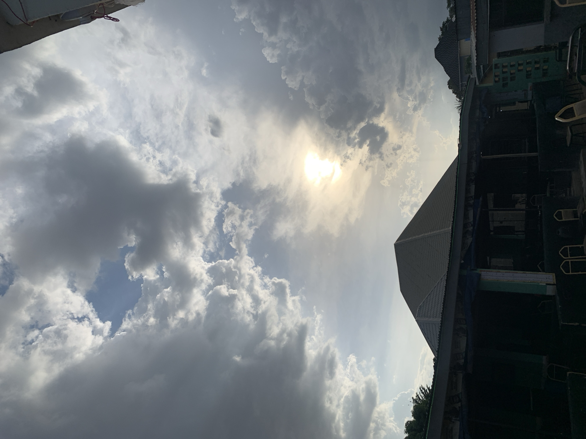 The image shows a sky with a mix of dark, heavy clouds and patches of clear blue, obscuring the sun. In the bottom right corner is part of a building, showing rooftops and sections of walls, giving context to the location in Kabong, Nigeria.