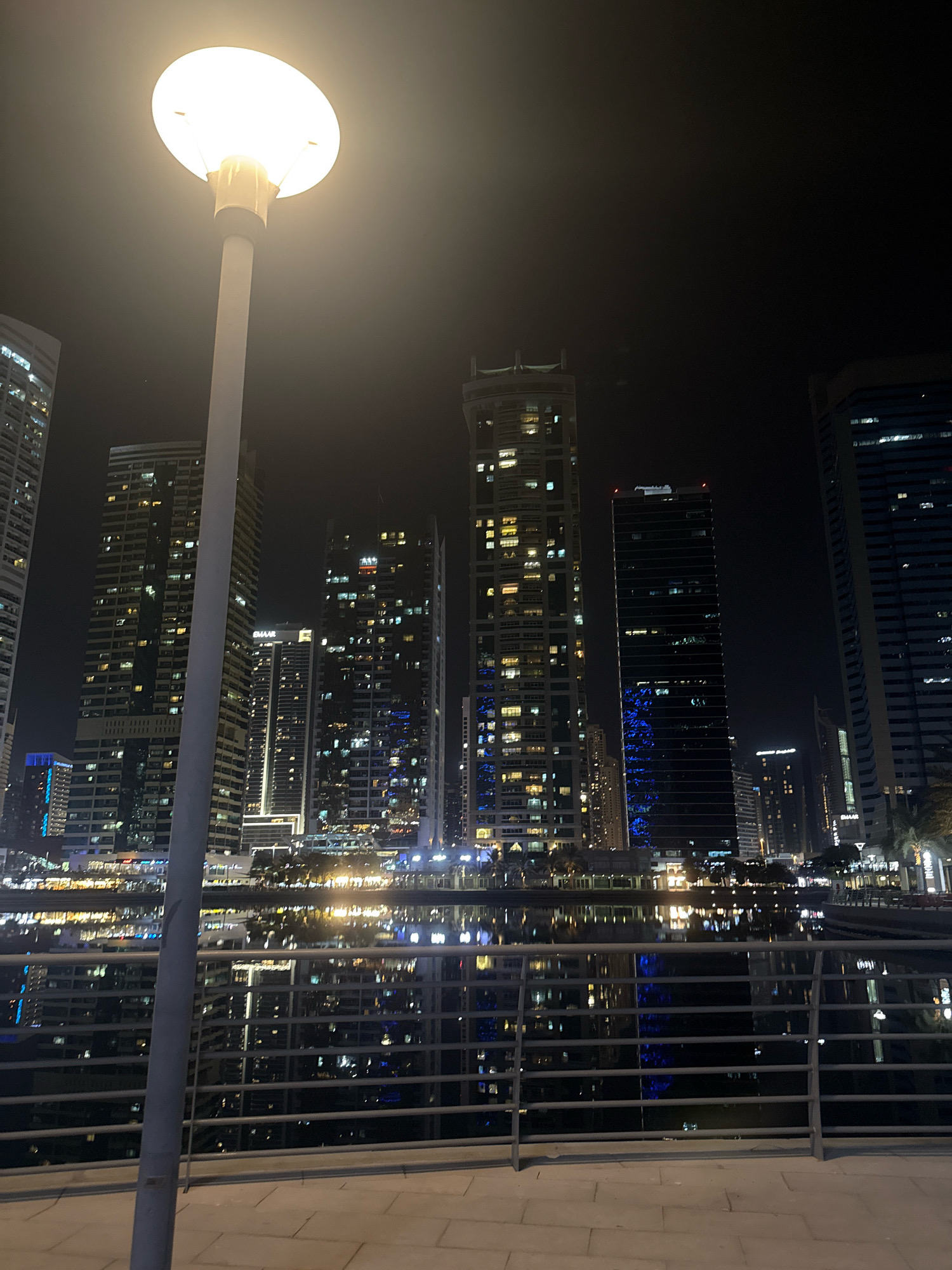 The image shows a night view of the Dubai Marina, a popular waterfront district in Dubai, United Arab Emirates. A tall lamppost with a bright light dominates the foreground on the left, casting a glow across the scene. Behind it, a body of water, likely the marina canal, reflects the lights of numerous skyscrapers that line its banks. These buildings are illuminated with a variety of lights, creating a vibrant and modern cityscape. The architecture is characterized by sleek, contemporary designs with many windows. A metal railing and a paved walkway are visible in the lower portion of the image, suggesting a promenade or viewing area. The sky is dark, emphasizing the illuminated urban landscape. There are no people visible in the frame.