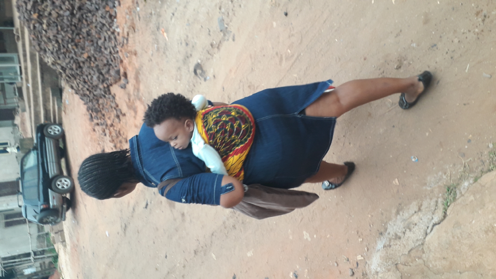 A woman is walking on a dirt surface in what appears to be a residential area in Obosi, Nigeria. The woman carries a baby on her back, secured with a colorful patterned cloth. She is wearing a denim dress and dark flats. A brown bag hangs from her shoulder. The area appears to be partially developed with a building and a dark-colored car visible in the background. There is also a pile of rocks or debris near the building. The ground has a mix of dirt and small patches of vegetation.