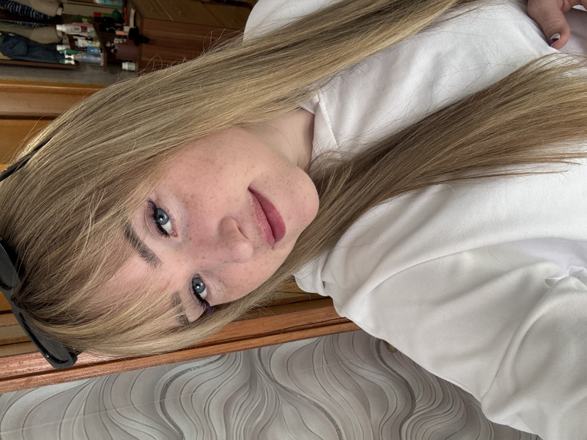 A fair-skinned woman with blonde hair and blue eyes is pictured. She has freckles on her face and is wearing lipstick. To her left are black sunglasses. She is wearing a white hoodie. The location could possibly be in Марковский, Russia. The background consists of wooden furniture and wallpaper with a wave pattern.