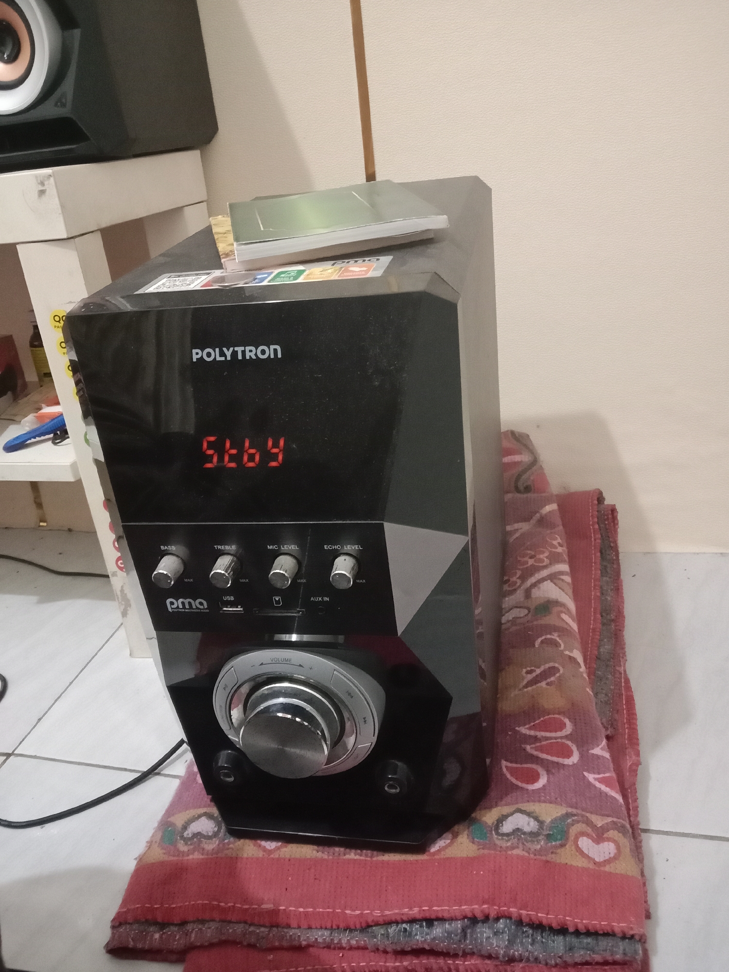 The image shows a black Polytron speaker with a digital display showing "5t64". There are several control knobs labeled "BASS", "TREBLE", "MIC LEVEL", and "ECHO LEVEL". Below these knobs are a "PMA" logo, a USB port, and an "AUX IN" port. A large silver volume dial is located at the bottom. The speaker is placed on top of a red and patterned cloth, which is itself on a tiled floor. In the background, a portion of another speaker and a white shelf are visible. The overall scene appears to be indoors, with an artificial light source. The location is identified as Brangsong, Indonesia.