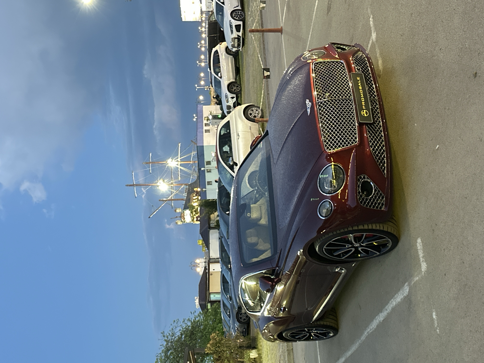 The image shows a burgundy Bentley parked in a parking lot in Varna, Bulgaria. The parking lot also contains several white and dark-colored vehicles parked in a row. A sailboat-shaped structure is visible in the background. The sky is blue with some clouds, suggesting it is dusk or dawn. A Promosale tag hangs from the car's grill. Street lights illuminate the scene. The parking lot is marked with white lines.