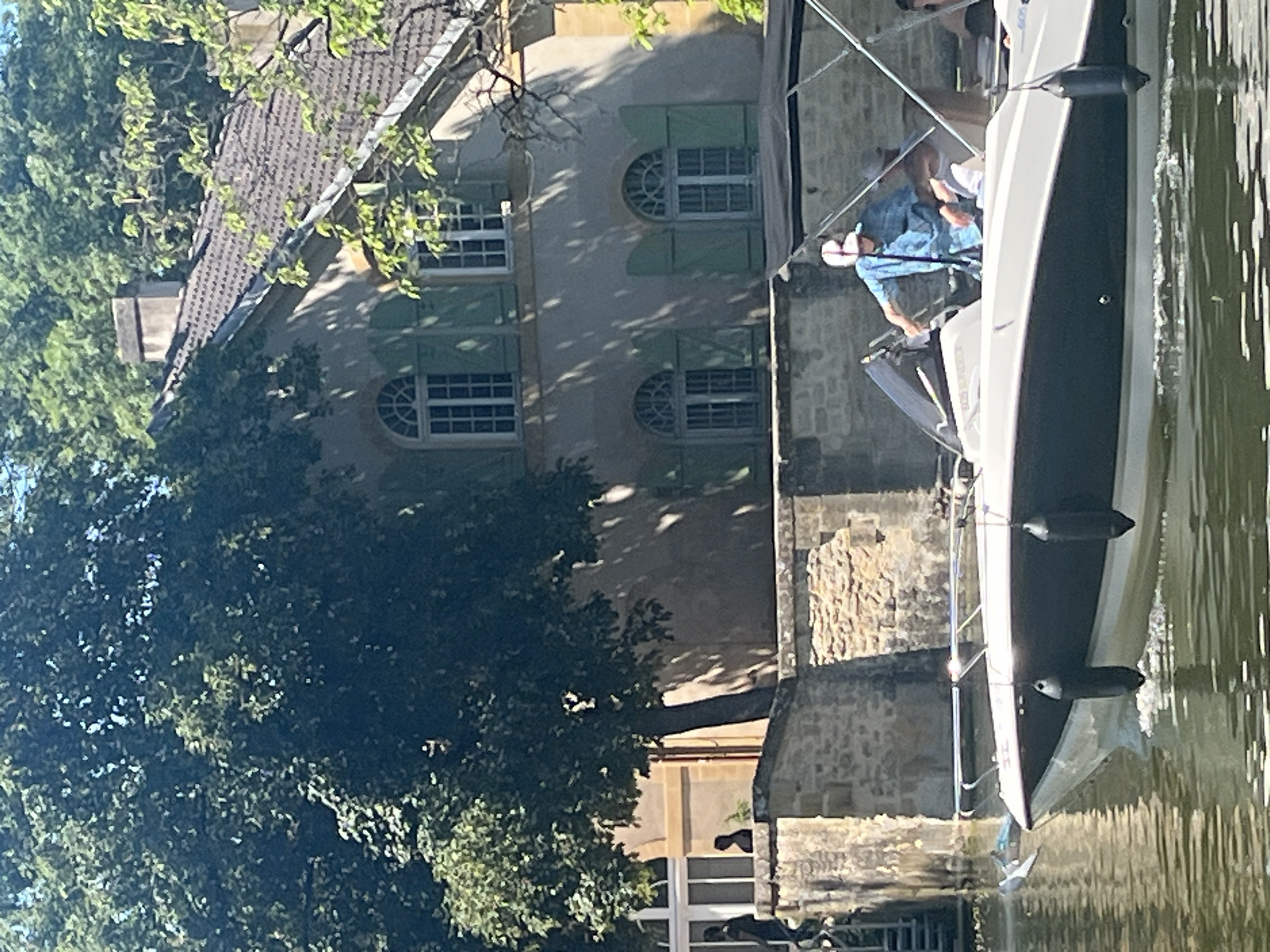 The image depicts a motorboat on the Moselle River in Metz, France, with a man operating it. The boat is positioned close to the riverbank where a stone building with a light-colored facade and green shutters is located. The building appears to be two stories high and has a tiled roof. A large tree with dense foliage partially obscures the view of the building. The river reflects sunlight, and the overall setting suggests a sunny day.