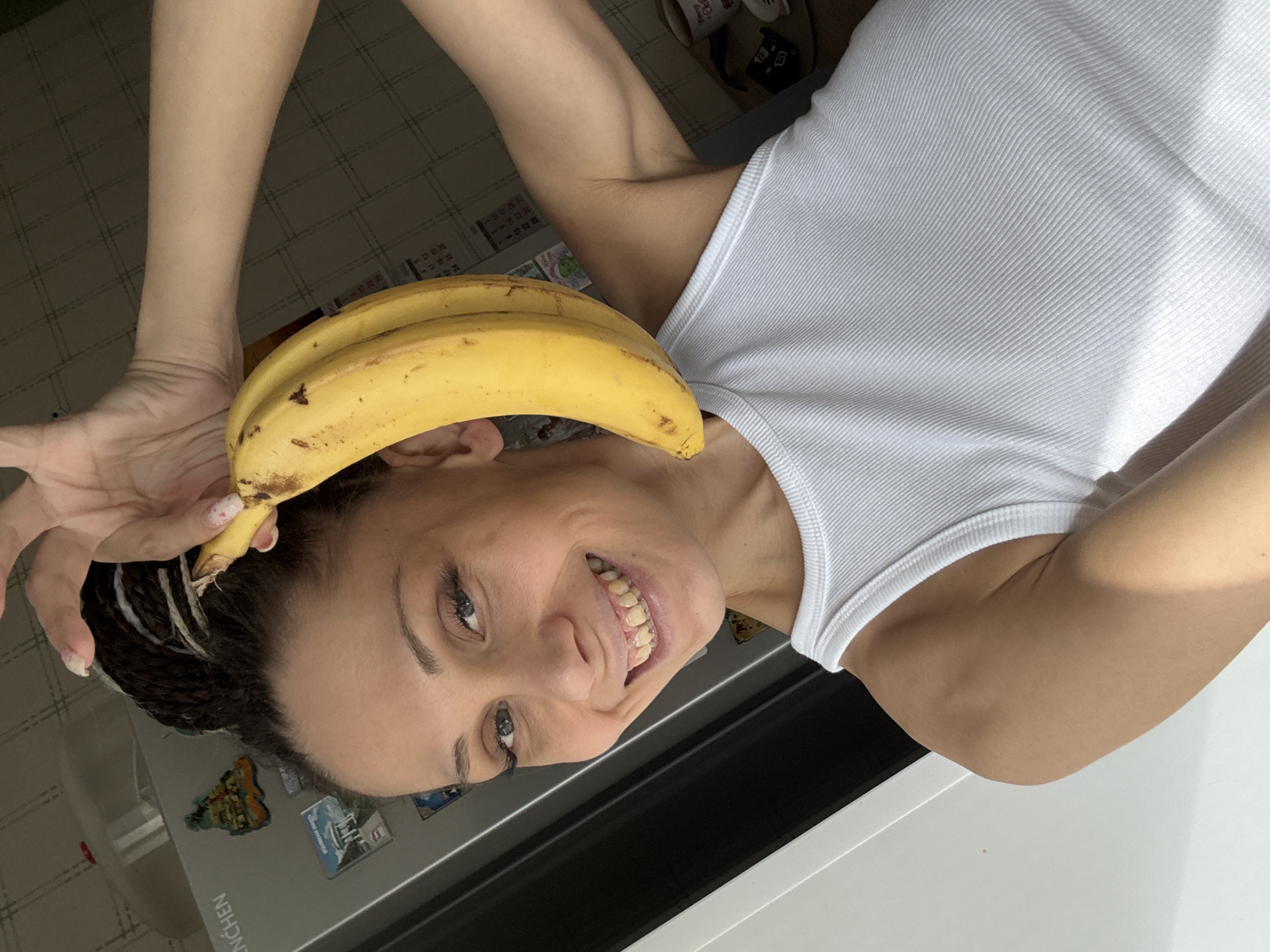 A woman with braided hair and a white tank top holds two bananas on top of her head. She is smiling and looking at the camera. She is indoors in a kitchen next to a white appliance with magnets on it. The location is Unknown, Unknown.