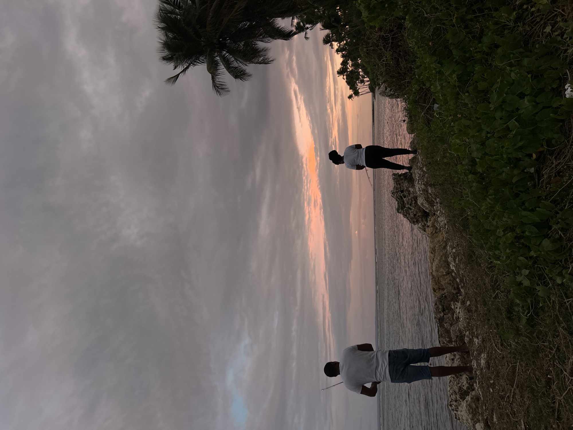 Two people are silhouetted on the coast at sunset. One person is wearing a white shirt and dark pants and holding a fishing pole. The other person is wearing a white shirt and shorts and is also holding a fishing pole. The coastline is rocky with some vegetation. A palm tree is visible in the background. The sky is cloudy with a pink and orange sunset. The location may be Sainte-Anne, France.