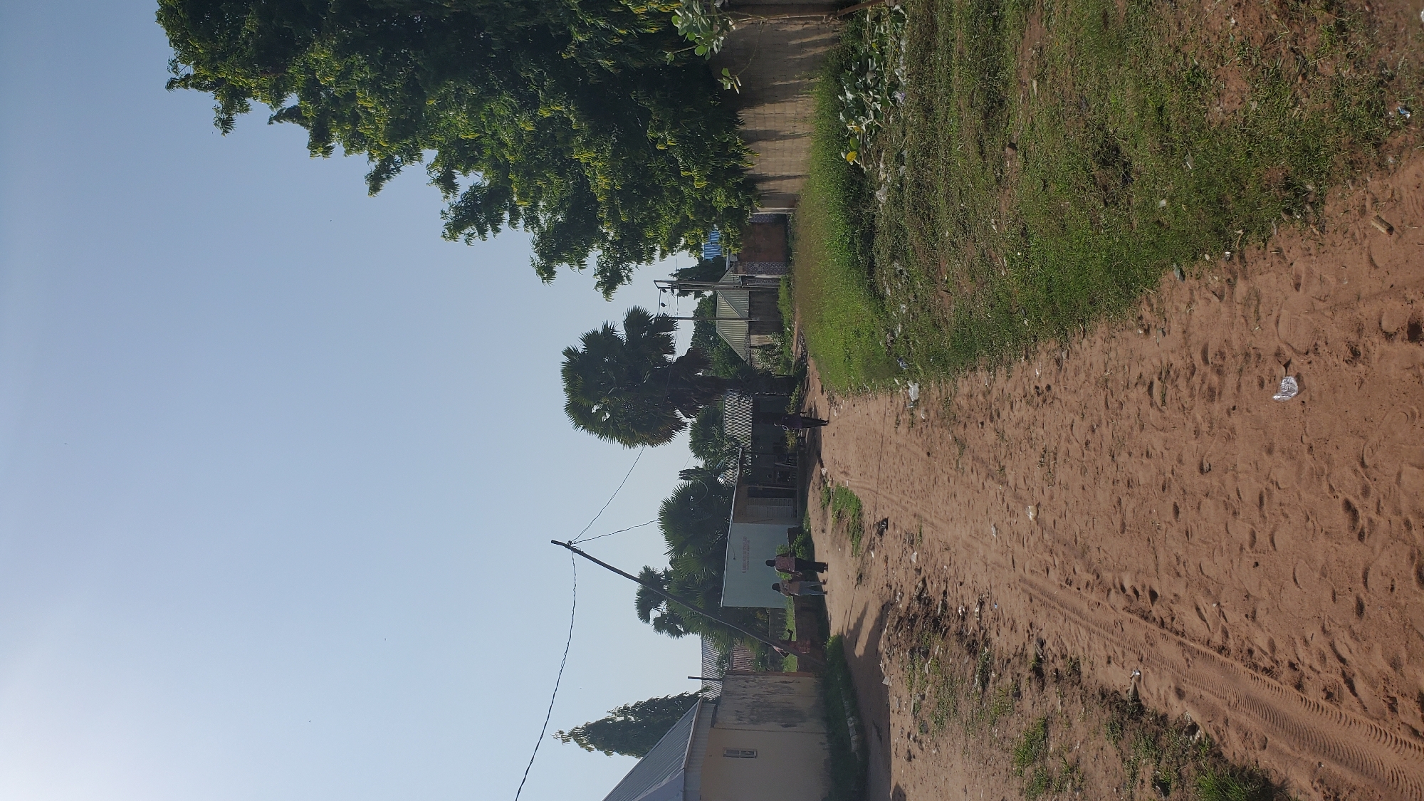 The image depicts a dirt road in a village setting, likely within the city of Jalingo, Nigeria. The road is uneven and shows tire tracks and footprints, with scattered litter along its edges. On the left side of the road, there are several buildings with corrugated metal roofs, some appearing to be houses or small shops. A tall, slanted utility pole with power lines runs across the scene. In the middle ground, a few people are walking on the road, including two individuals close together and one person further down the path. Palm trees and other lush green vegetation are visible throughout the image, with a dense tree line and a stone wall on the right side. The sky is clear and bright blue.