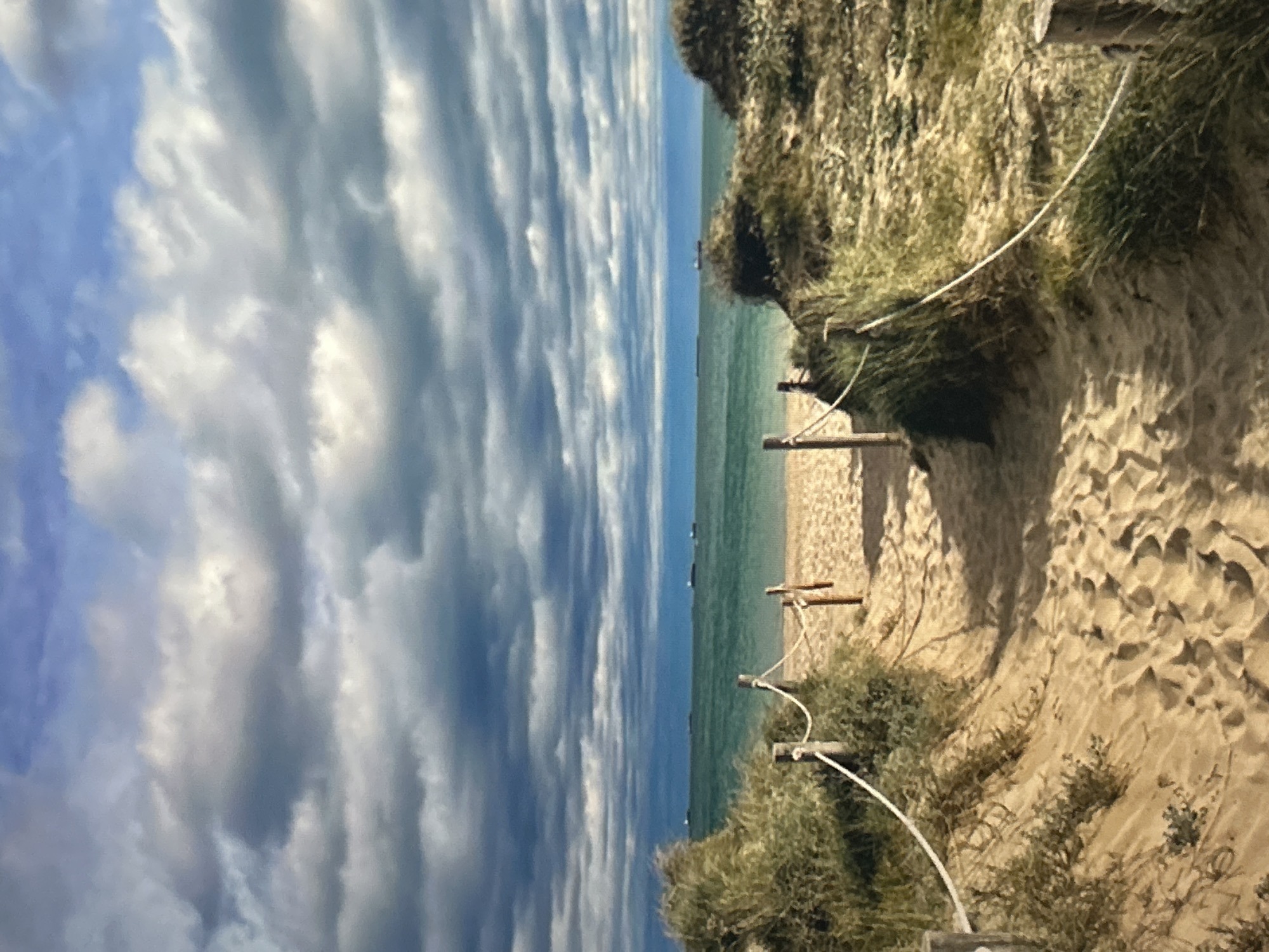The image depicts a serene coastal landscape, offering a view of a sandy path leading down to a beach with the sea and sky beyond. The perspective suggests the original capture might have been in portrait mode and then rotated.
The top portion of the frame is dominated by a vast sky filled with elongated, stripy clouds in shades of white and grey, interspersed with patches of blue. These clouds appear to stretch horizontally across the sky, creating a sense of movement or a unique atmospheric condition.
Below the sky lies the tranquil turquoise sea, which extends to the horizon. The water appears calm, and in the distance, a few faint, dark shapes are visible on the water, possibly indicating distant boats or ships.
To the right, a sandy dune or cliff rises, covered with dry, sparse green and brown vegetation, consistent with a coastal environment. A sandy path curves downwards along this dune, leading towards the water's edge. This path is bordered by several wooden posts connected by a white rope, acting as a guide or barrier. The sand on the path and dune exhibits textured patterns.
There are no people or animals visible in the scene. The lighting suggests it is daytime, with a soft, natural illumination highlighting the textures of the sand and clouds. The overall mood is peaceful and natural, showcasing a picturesque beach access point. No text is visible in the image.
