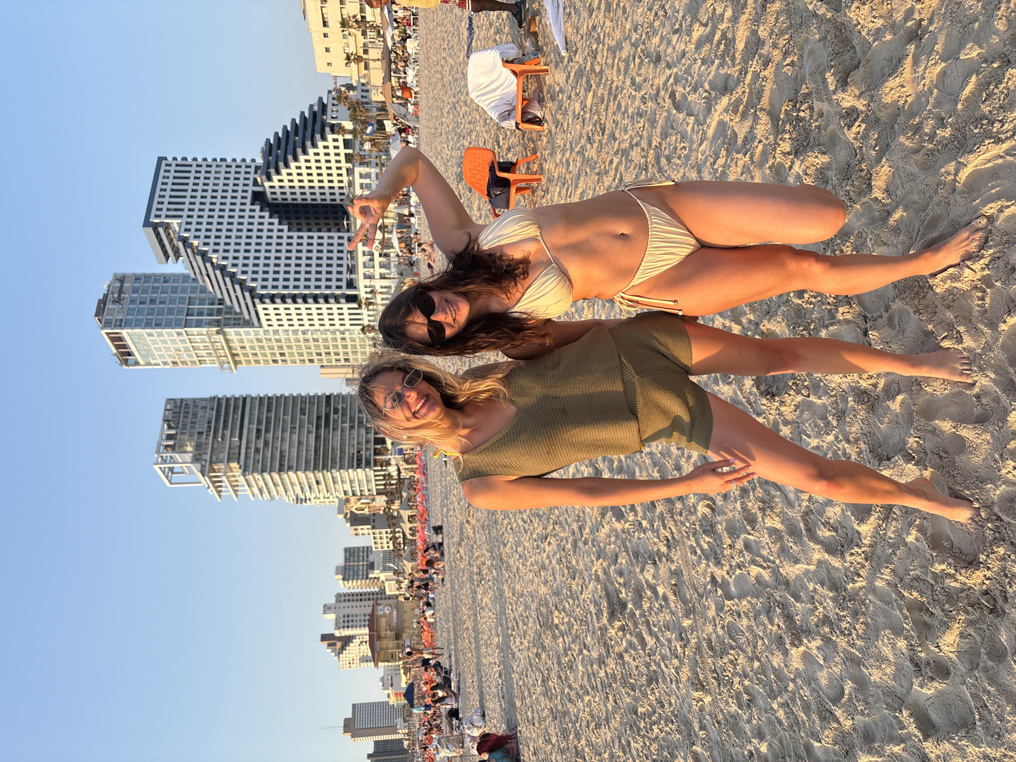 The image captures two young adult women posing on a sandy beach in Tel-Aviv, Israel, during the late afternoon.
The woman on the left has blonde hair and wears sunglasses. She is dressed in an olive-green knitted sleeveless top and matching shorts, standing with her weight on her right leg and her left leg slightly bent. Her left arm hangs casually, and her right hand rests on her right thigh. She smiles directly at the camera.
The woman on the right has darker hair and wears sunglasses. She is dressed in a light yellow string bikini. She stands with her right arm raised, forming a peace sign with her fingers, and her left arm slightly bent. Her knees are slightly bent, and she also smiles directly at the camera. Both women are barefoot in the sand.
In the middle ground, numerous individuals are visible on the beach, some seated on orange and white lounge chairs, others standing. A lifeguard station is partially visible in the distant left.
The background features the distinctive skyline of Tel-Aviv. Prominently visible are several modern high-rise buildings. The building on the far left is a multi-story structure. To its right, a tall, light-colored skyscraper with a visible "Kempinski" sign on its upper facade identifies it as the Kempinski Hotel Tel Aviv. Adjacent to it on the right is a unique, multi-tiered white building with a distinctive stepped, angular design, which is part of the David Promenade Residences complex. Further right, other residential buildings and palm trees line the beach. The sky is clear and light blue, indicating fair weather. The low sun casts a warm, golden light across the sand and illuminates the subjects from the side, suggesting the approach of sunset.
